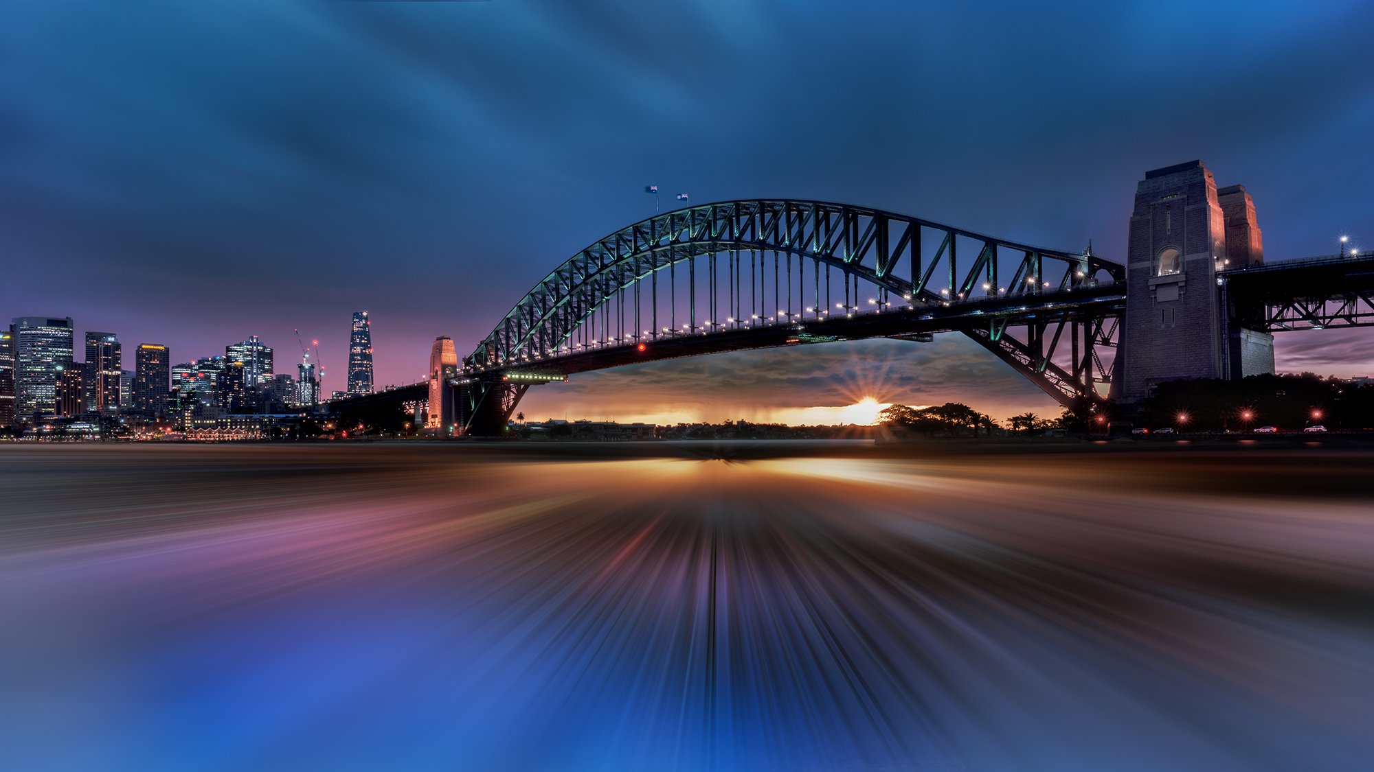 Nighttime view of the Sydney Harbour Bridge with city skyline in the background, sunset on the horizon, and water reflecting lights in the foreground.