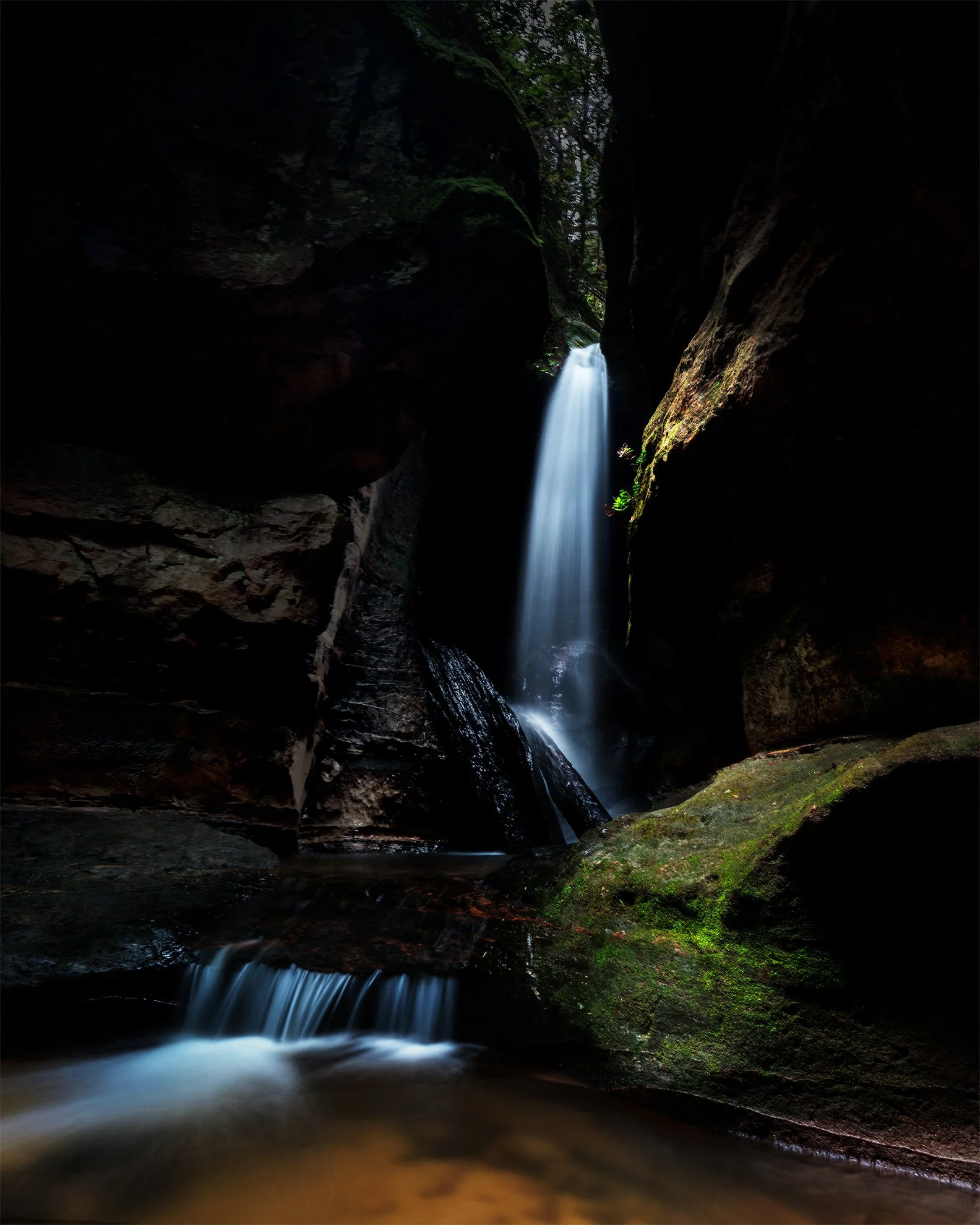 A narrow waterfall flowing through a dark, rocky canyon with moss-covered rocks.
