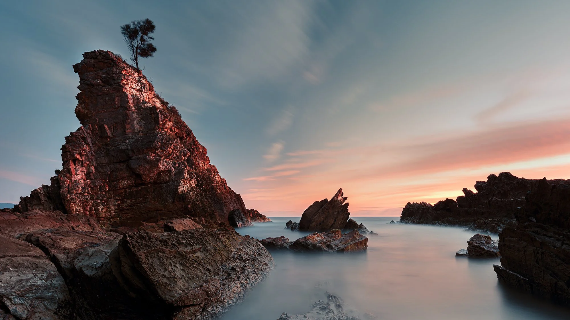 Sunset over a rocky coastline with large cliffs and a tree on top, calm water with rocks, and a sky with pink and blue hues.
