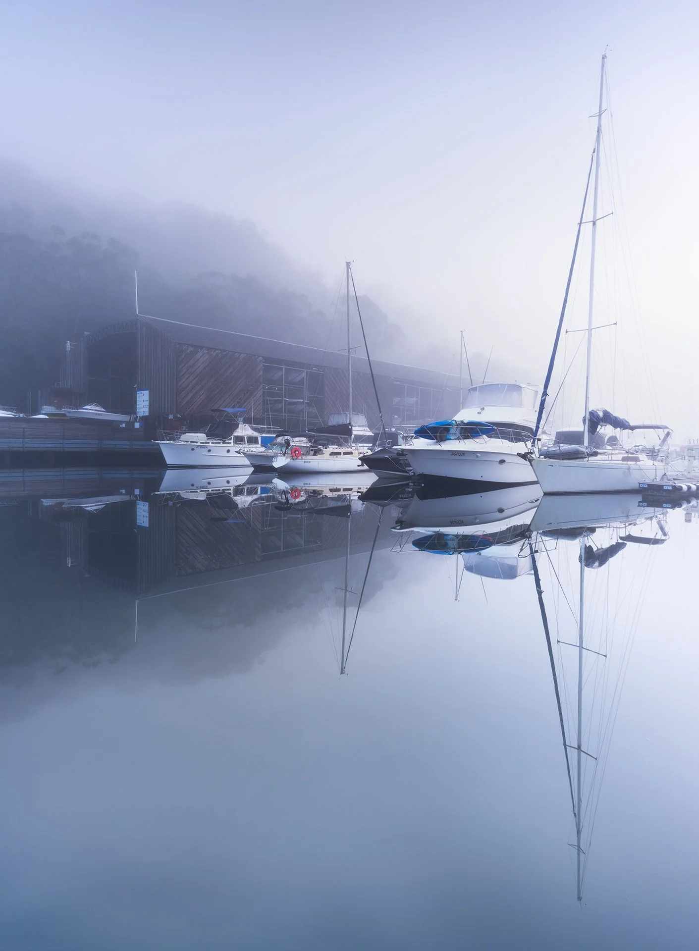 Marina with boats docked, foggy mountain background, reflections on calm water.