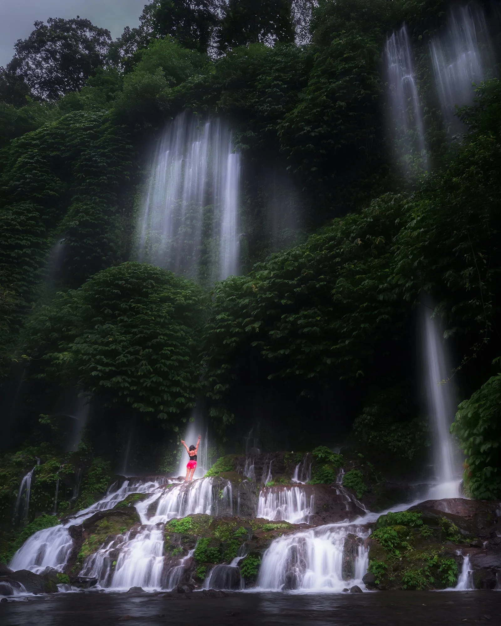 Person in pink shorts and black top standing on rocks under a multi-tiered waterfall in a lush green forest.