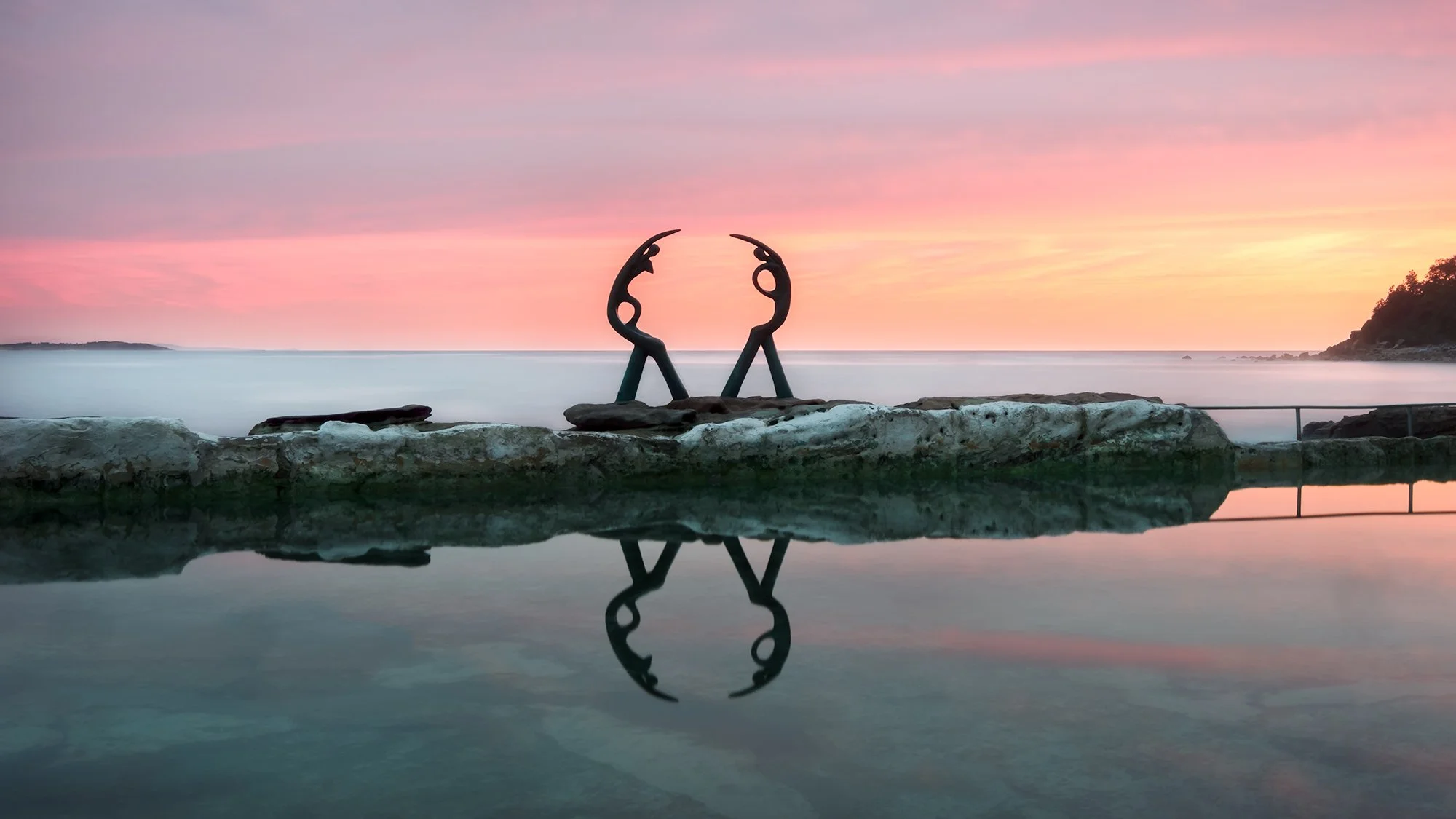 A sculpture of two abstract figures holding hands, located on a rocky outcrop by a calm body of water during sunset, with reflections visible on the water's surface.