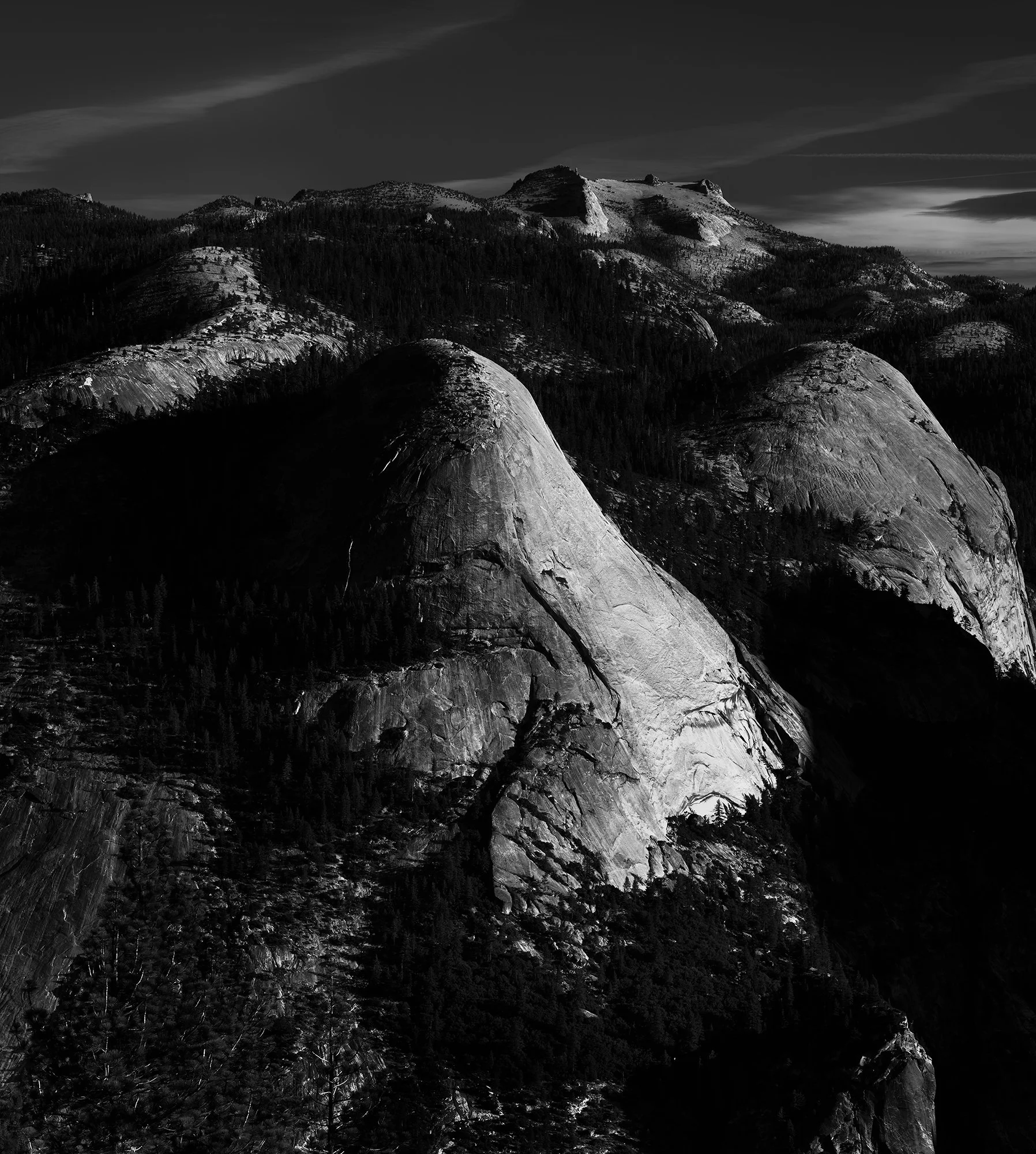 Black and white photo of rugged mountain landscape with large granite domes, steep cliffs, and dense forest at the base.
