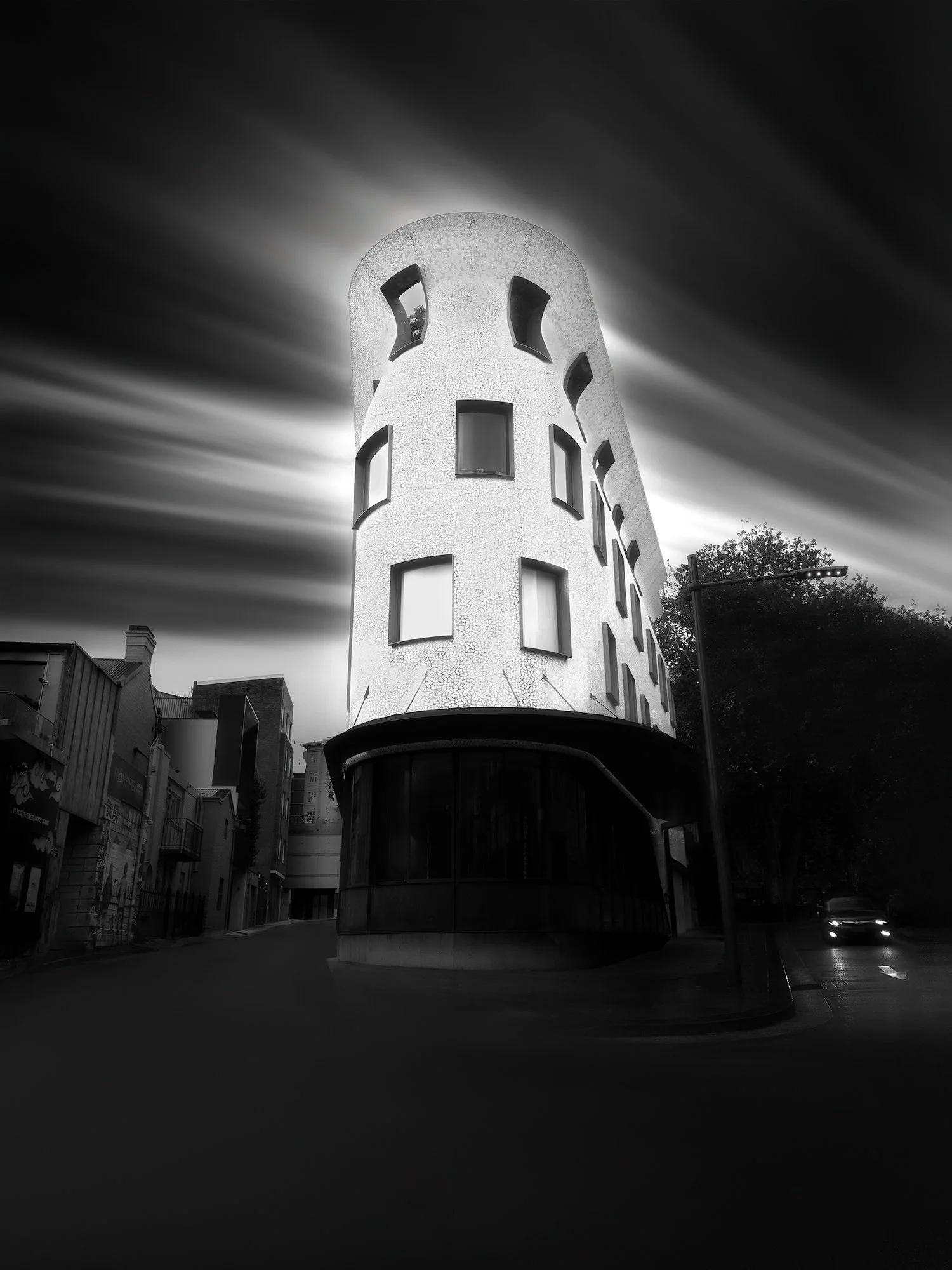 Nighttime black and white photo of a tall, rounded, multi-story building with numerous small, irregularly shaped windows, with a dramatic sky and motion blur clouds in the background.
