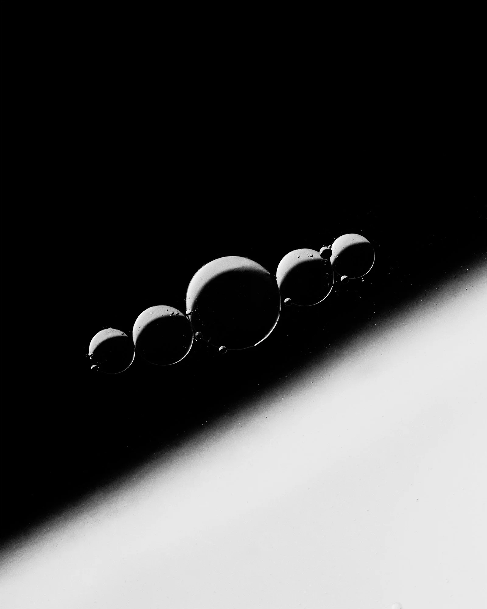 Black and white abstract image of bubbles or droplets aligned in a diagonal pattern, with a high contrast background divided diagonally into dark and light sections.