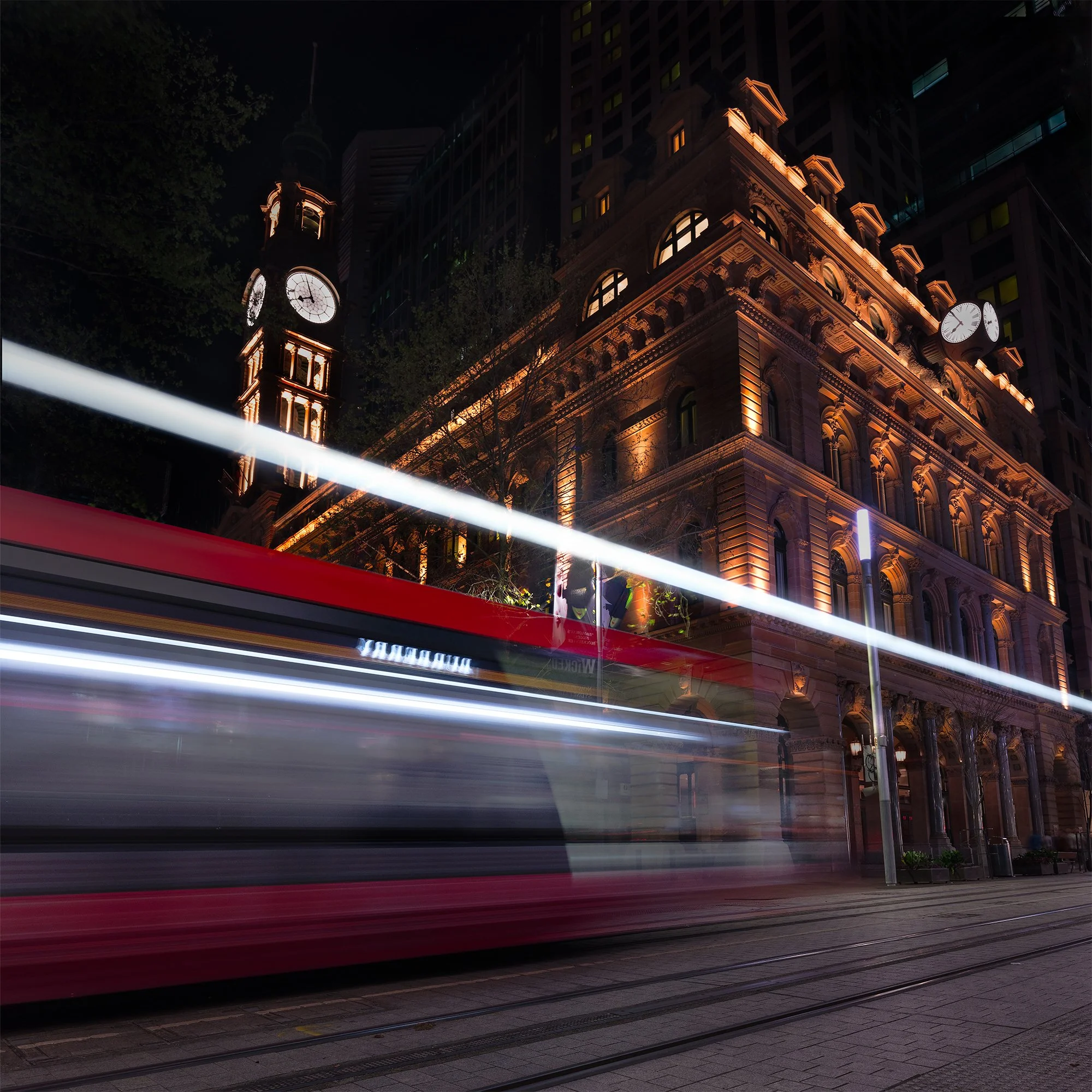 Night scene of a historic building with illuminated clock tower, light trails from passing streetcar, and urban streetlights.