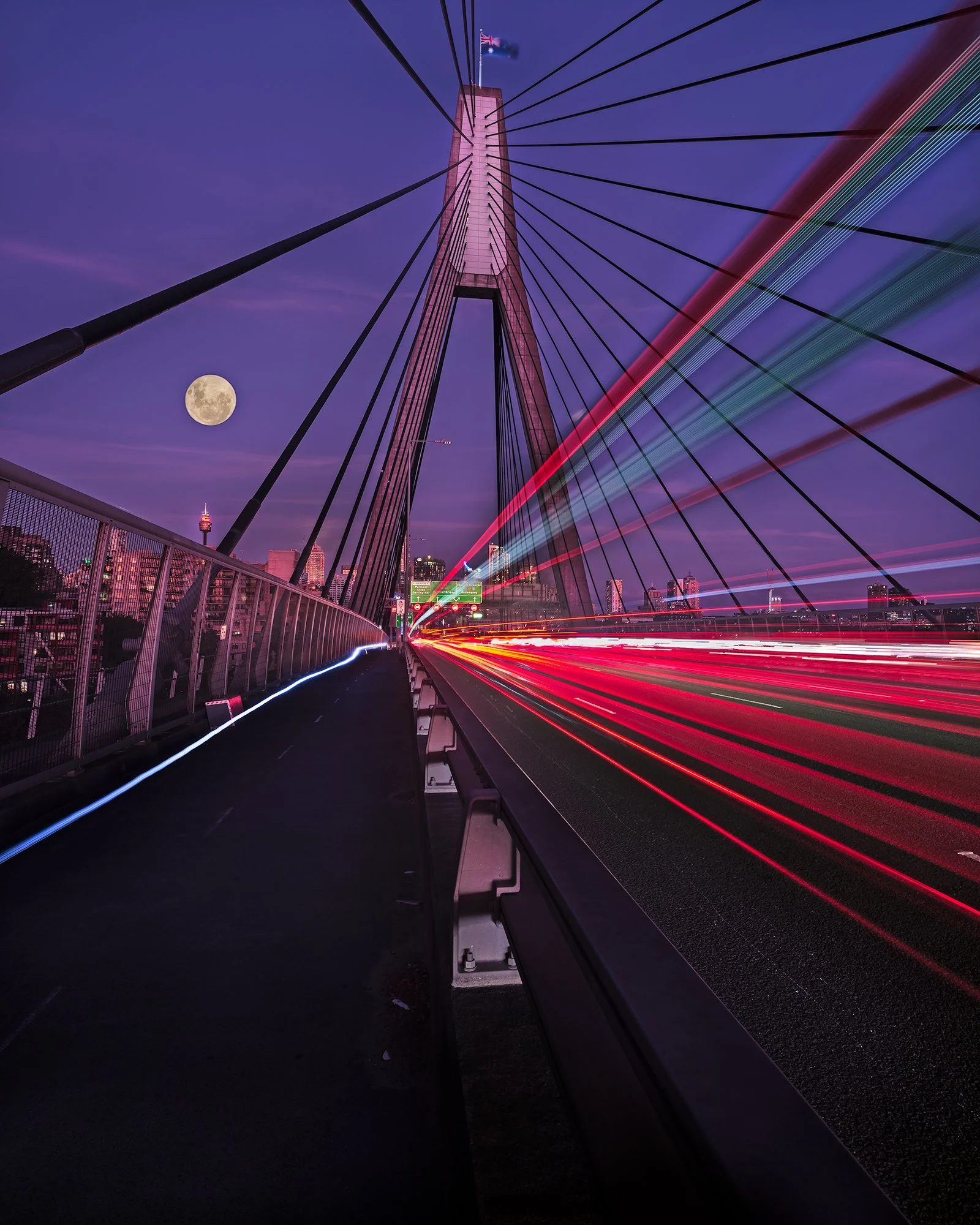 Long exposure photo of a bridge at night with streaks of vehicle lights, city buildings in the background, a bright full moon, and purple sky.