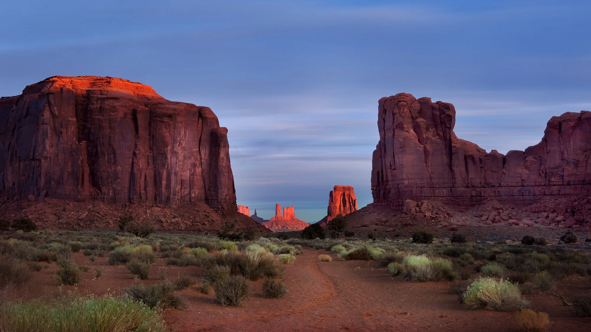 A desert landscape featuring large red rock formations and buttes under a sky with light clouds.