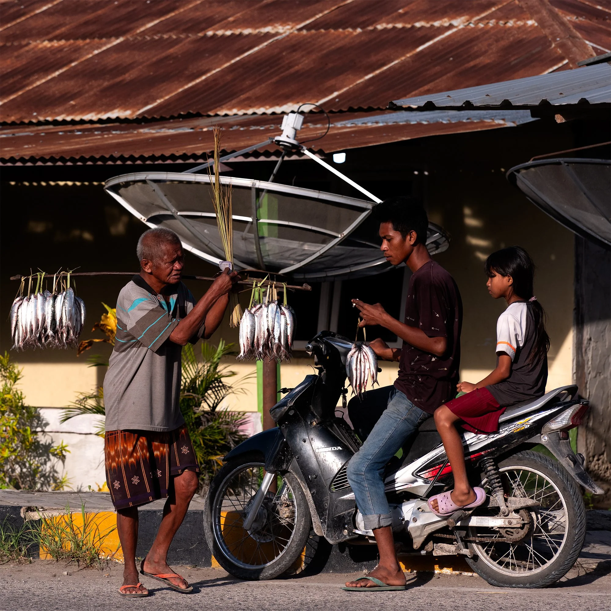 An elderly man selling fish hung on a stick to a young man and a girl on a motorcycle in a rural setting with satellite dishes overhead.