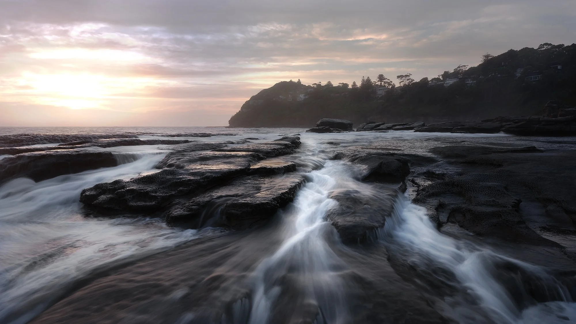 Sunset over rocky shoreline with waves flowing over black rocks, hills with trees and houses in the background.