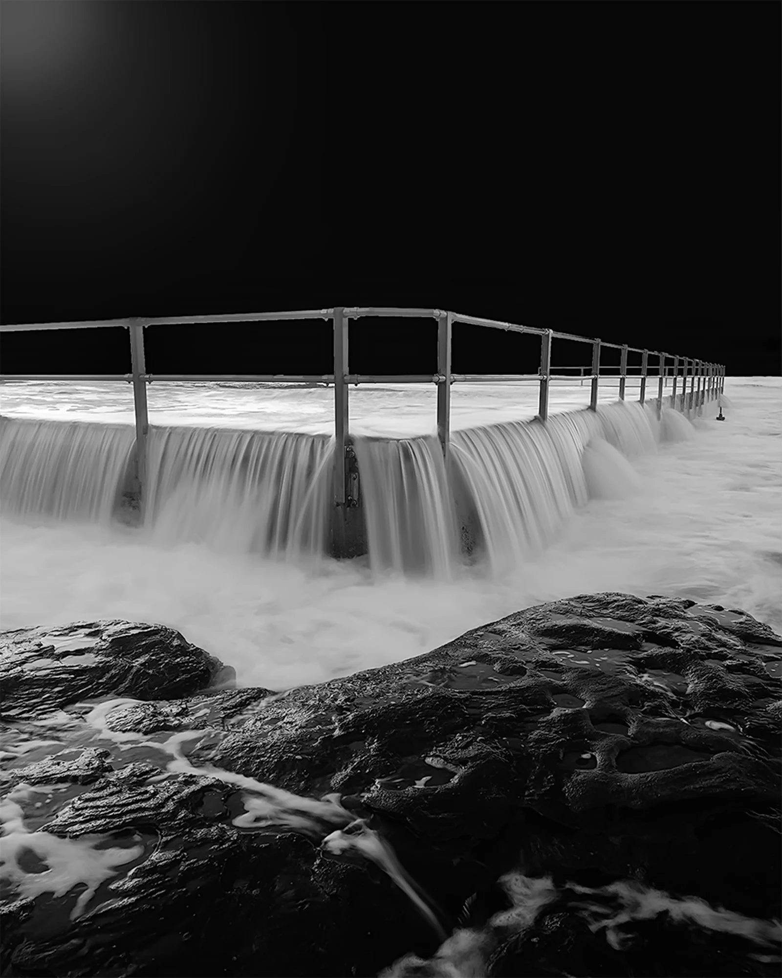 Black and white photo of a metal water filtration or drainage structure with water flowing over the edge, set against a dark sky, and rocks in the foreground.