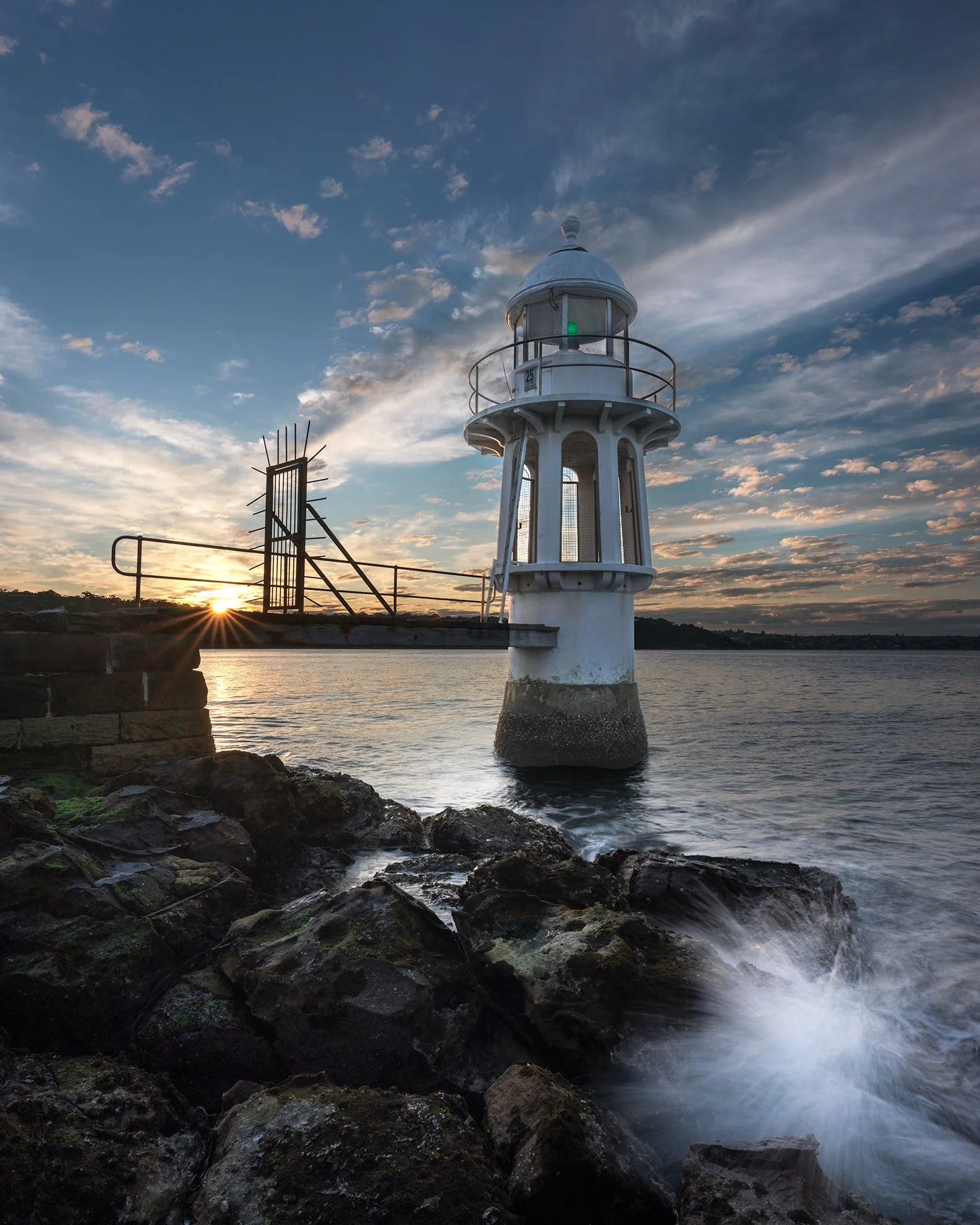 A lighthouse by the water at sunset, with rocks in the foreground and a partly cloudy sky.