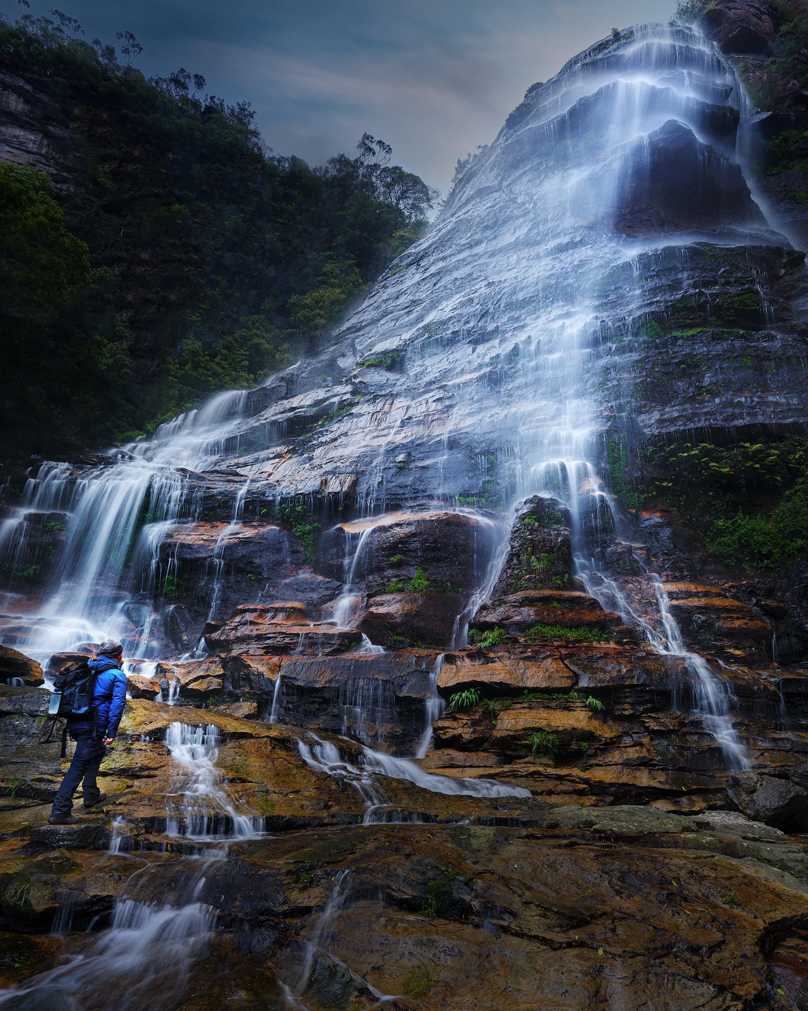 A person in outdoor gear standing on rocks and looking at a large waterfall cascading down a rocky cliff with greenery around, under a cloudy sky.