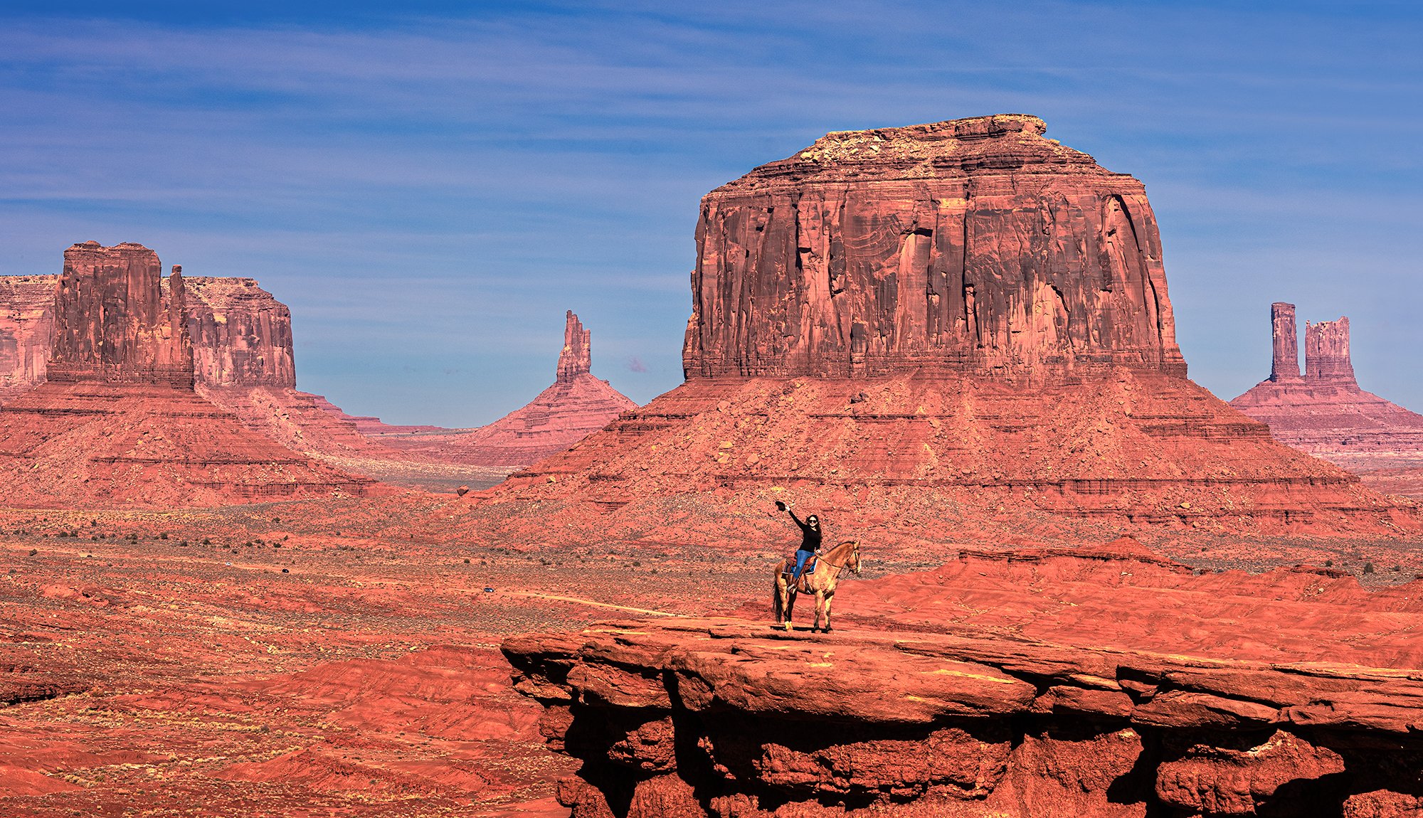 A person riding a horse on a rocky outcrop in the desert with large red rock formations in the background including buttes and mesas under a blue sky.