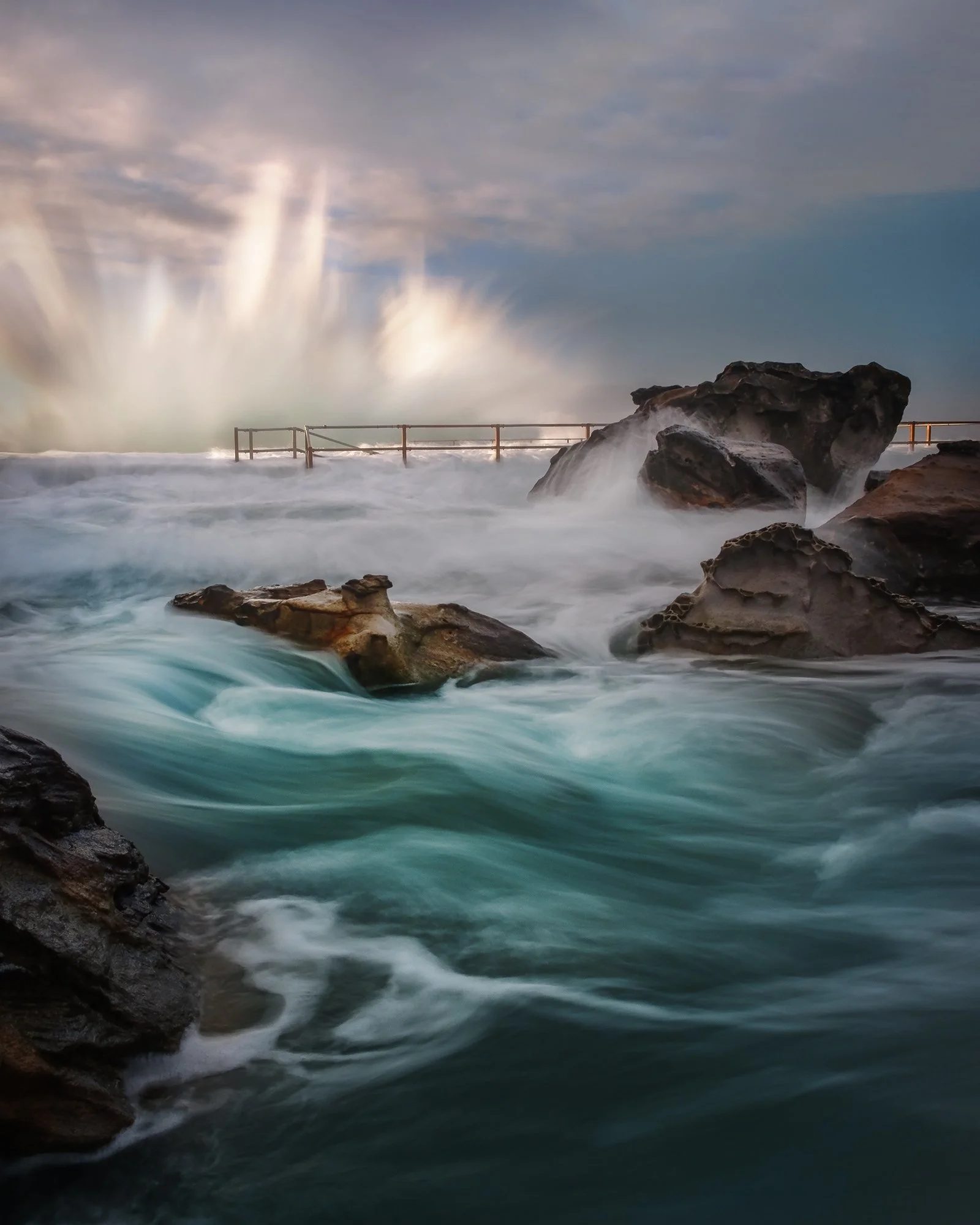 Waves crashing against rocks at a shoreline with a cloudy sky and a railing in the background.