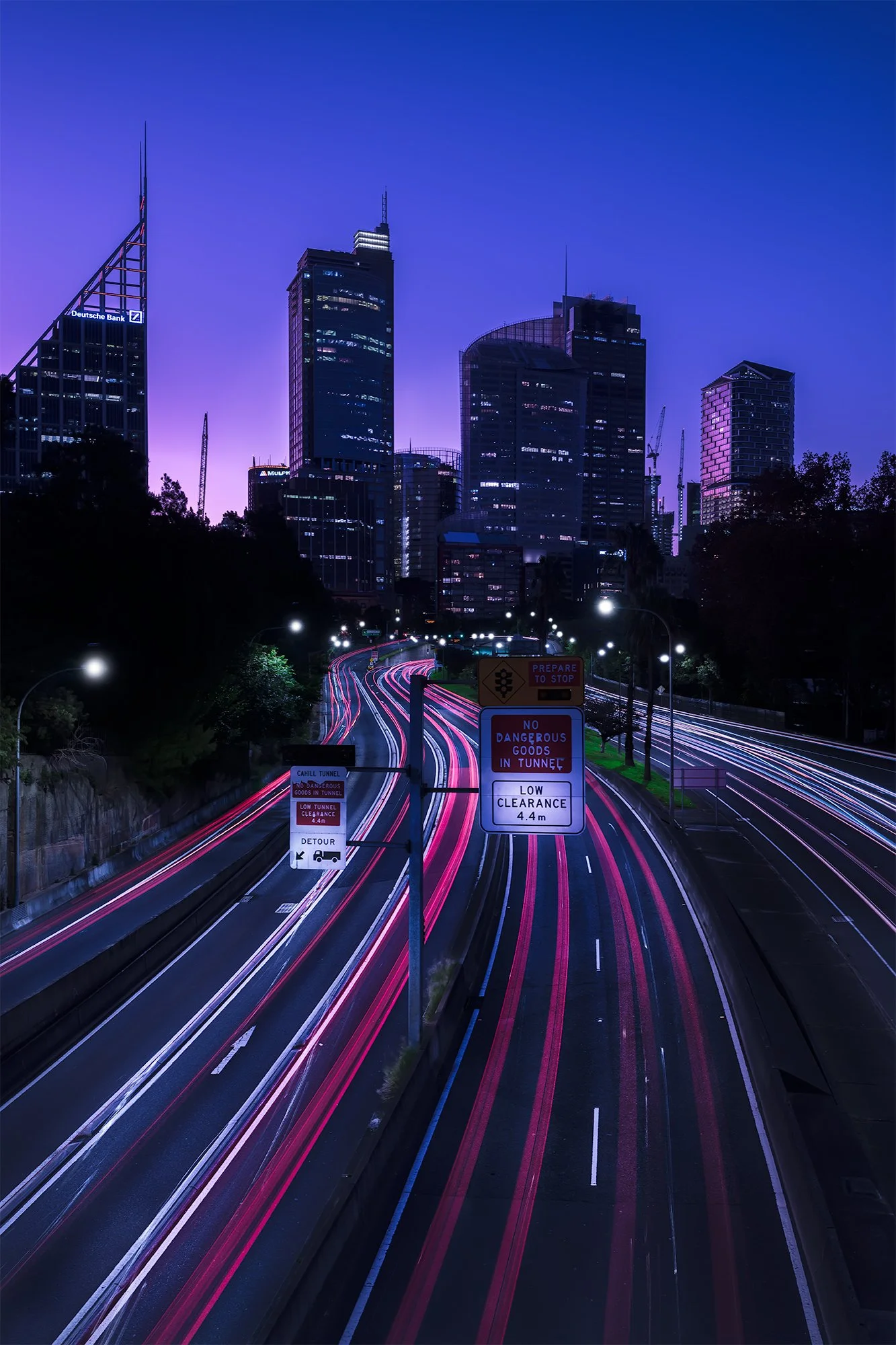 Nighttime cityscape with tall buildings and a highway with light trails from moving vehicles, illuminated signs, and a gradient purple to blue sky.