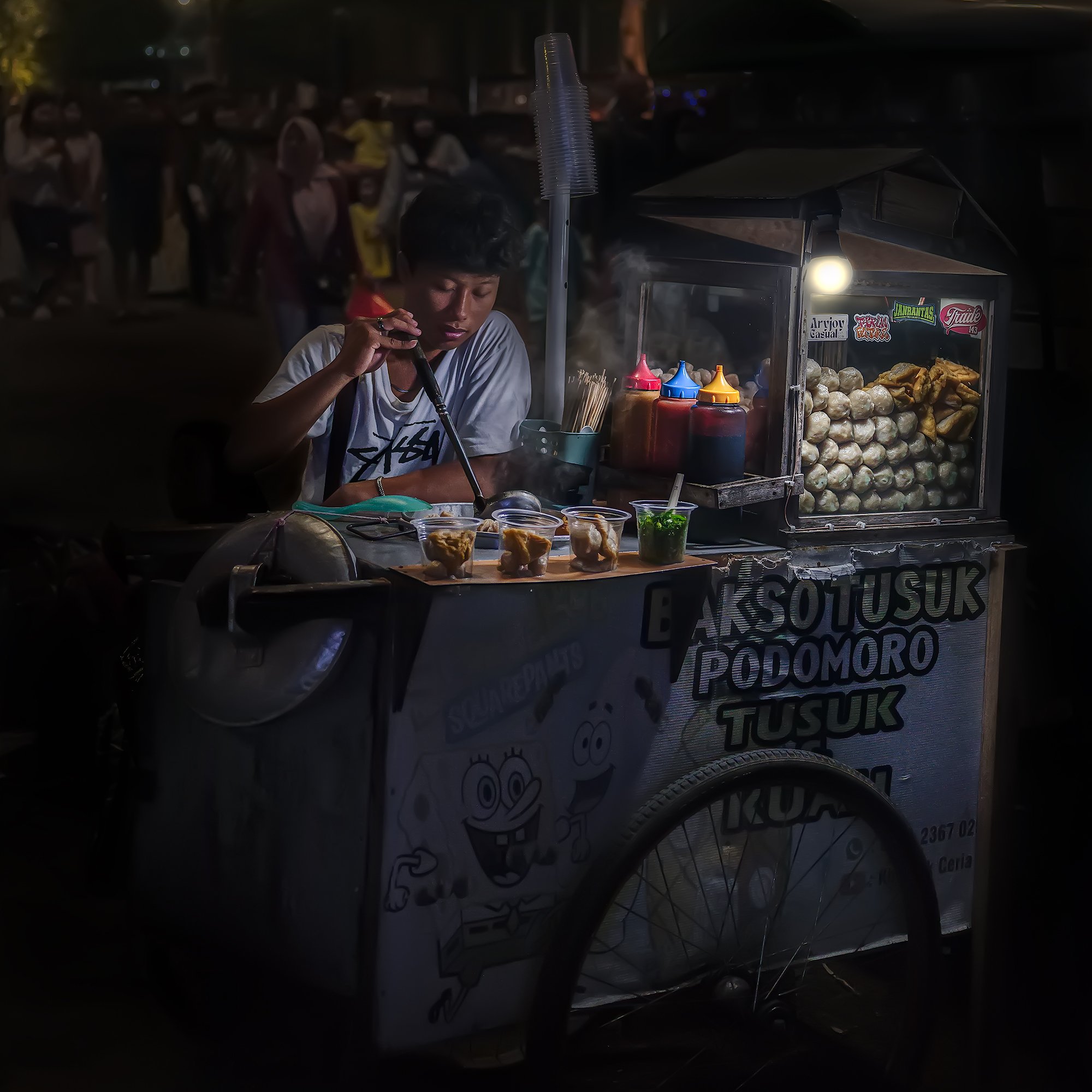 A young man preparing food on a street food cart at night, with various condiments and ingredients around him, and a crowd of people in the background.