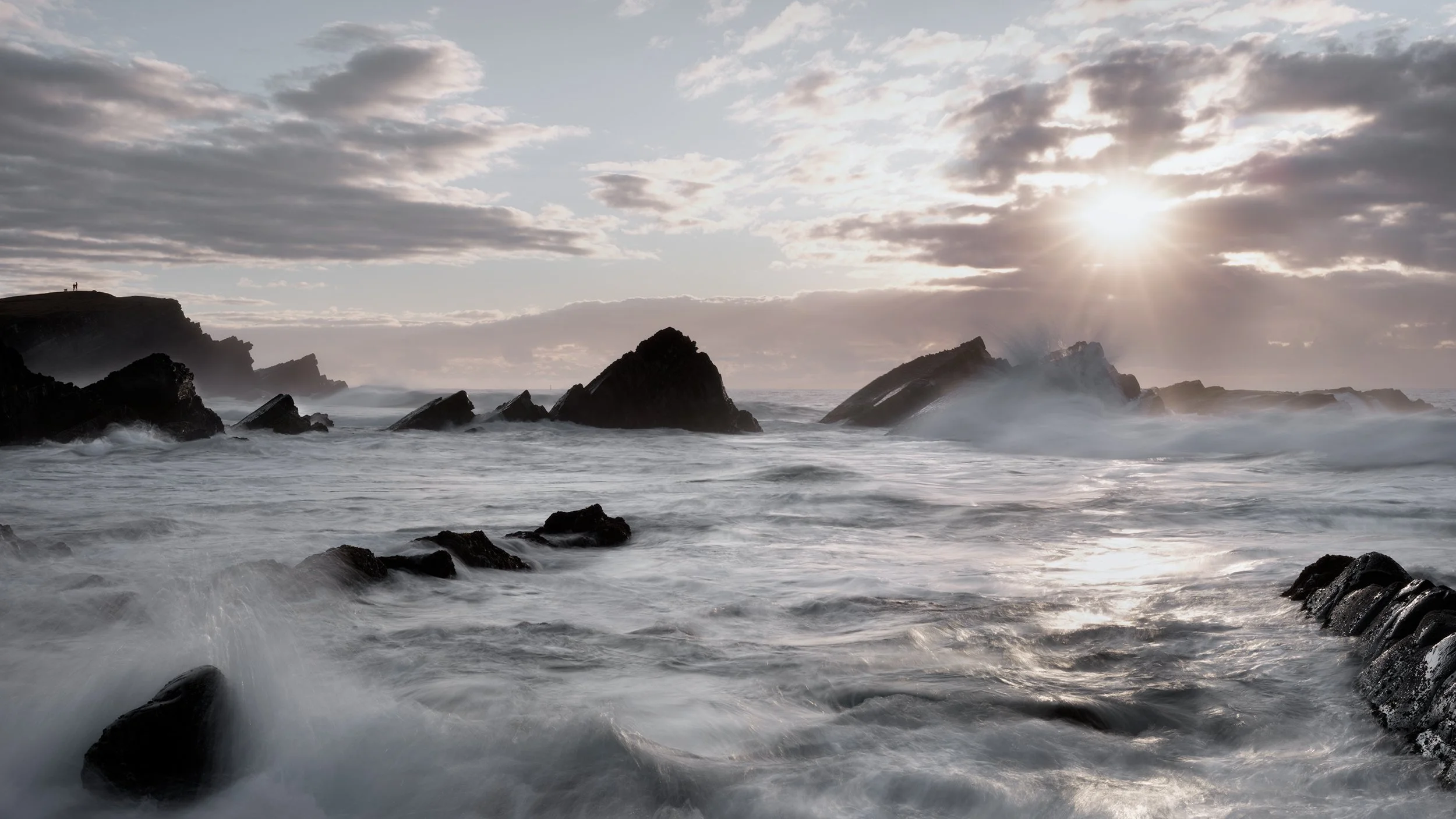 Sun shining over rocky coastline with waves crashing and a cloudy sky.
