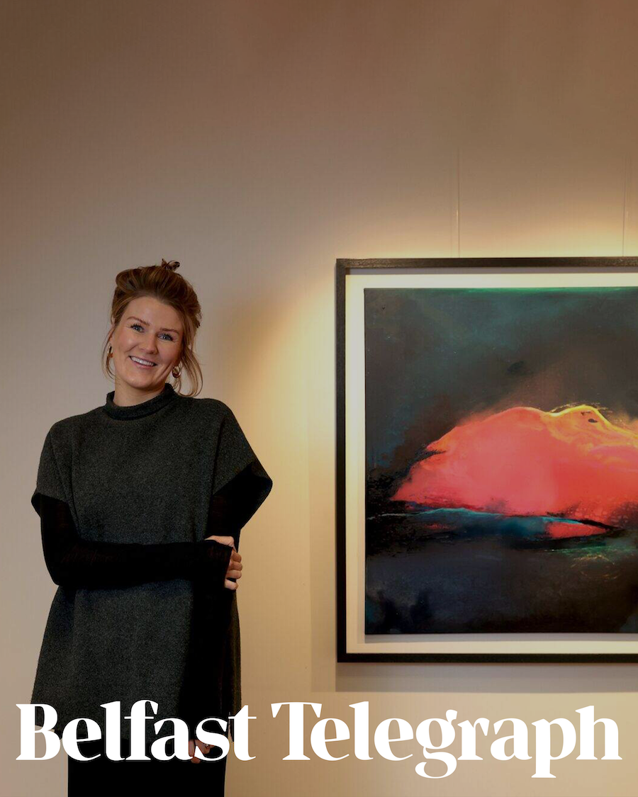 A woman with red hair smiling and standing next to an abstract painting with dark tones and bright orange and pink highlights. The woman is wearing a dark grey dress and is standing in front of a beige wall with lighting.