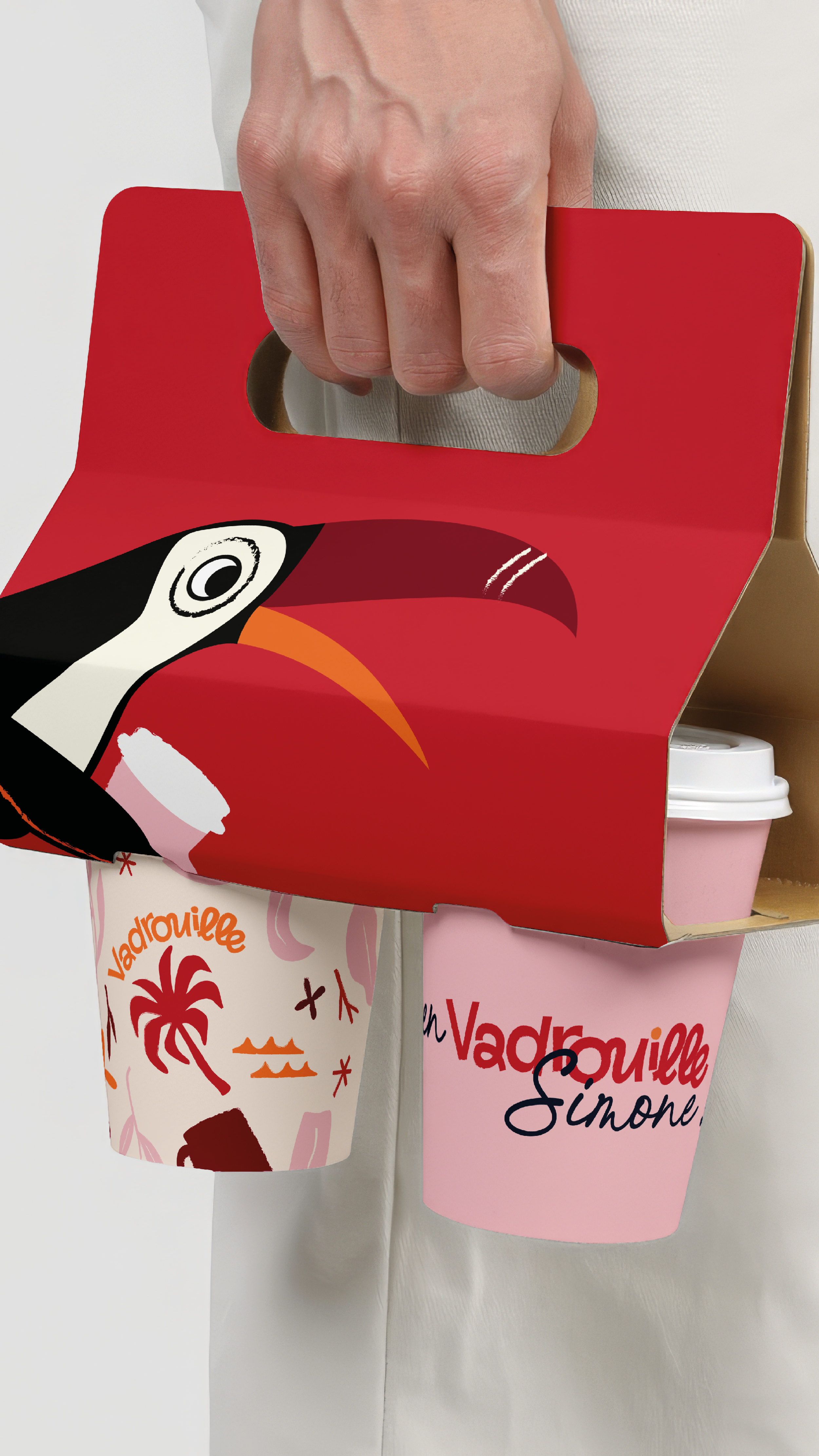 Une main tenant une boîte rouge ornée d'une illustration de toucan, contenant deux tasse à café. Identité Vadrouille !