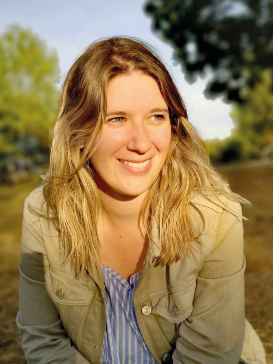 Jeune femme avec cheveux blonds, souriante, portant une veste beige et une chemise bleue à rayures, en plein air avec des arbres en arrière-plan.