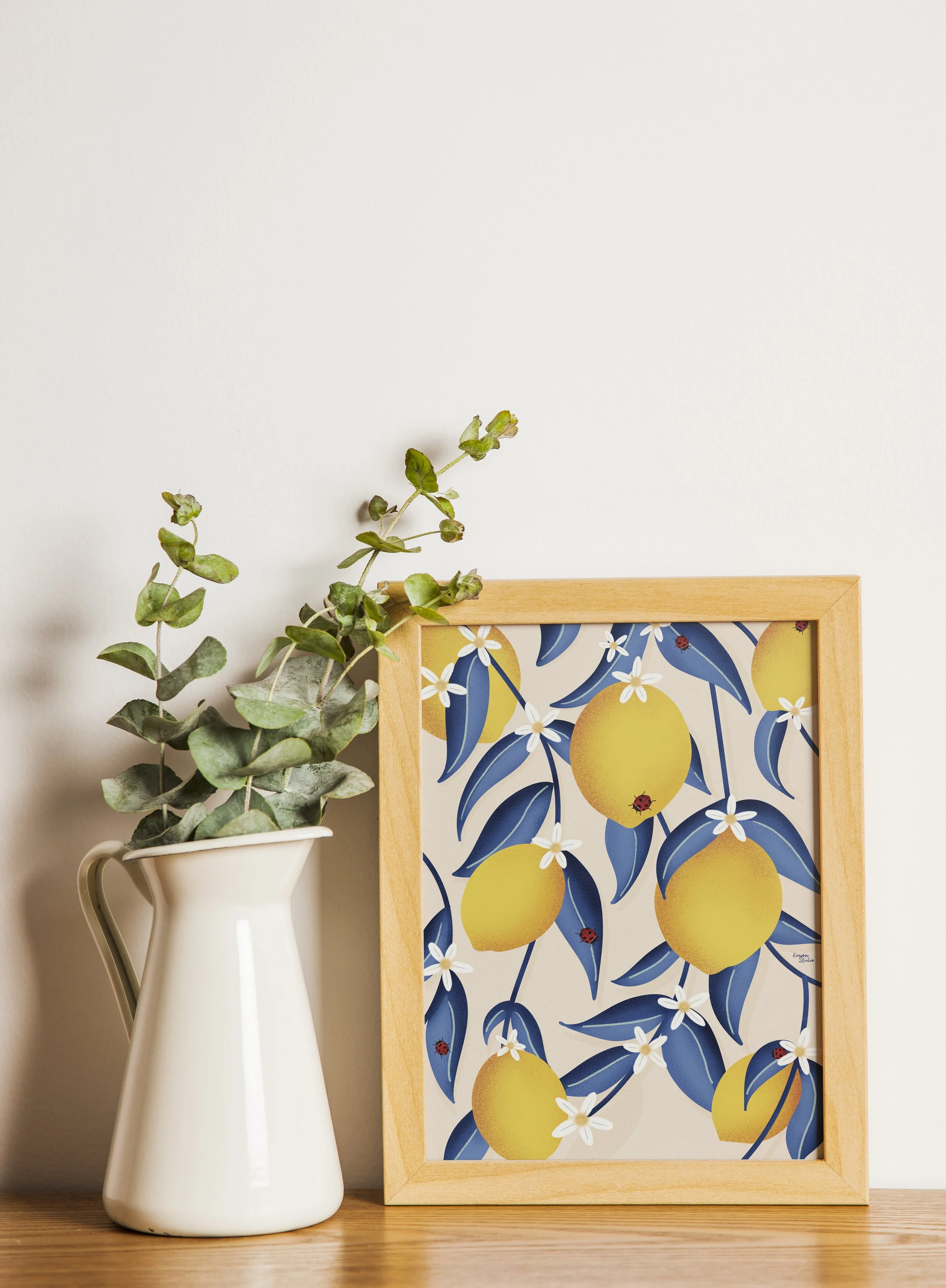Un vase blanc avec des branches de verdure vertes et grisées à côté d'une affiche encadrée représentant des citrons jaunes, des feuilles bleues, des petites fleurs blanches et des coccinelles.