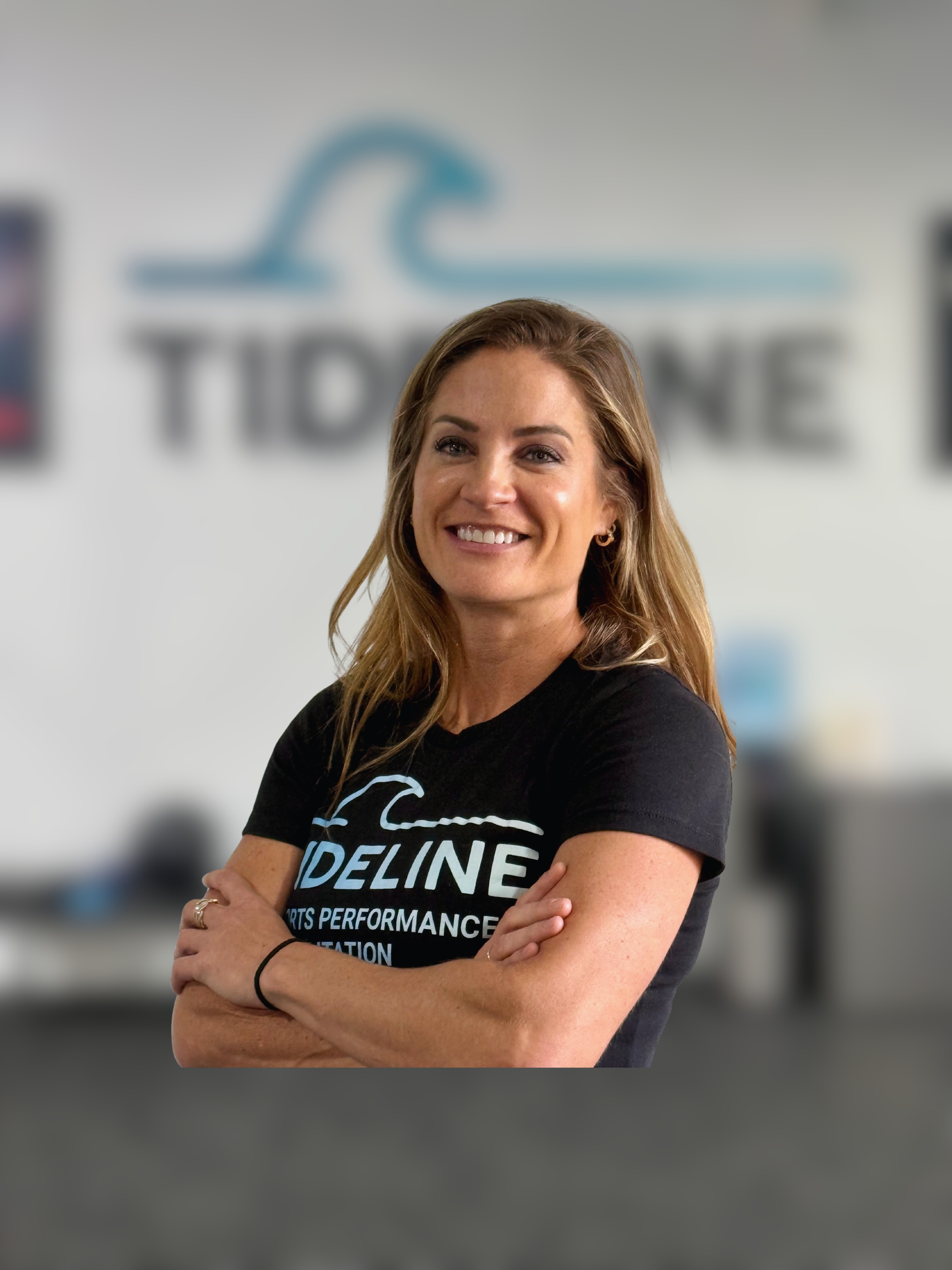 A woman smiling with crossed arms wearing a black t-shirt in front of a blurred logo of 'Tideline' on a wall.