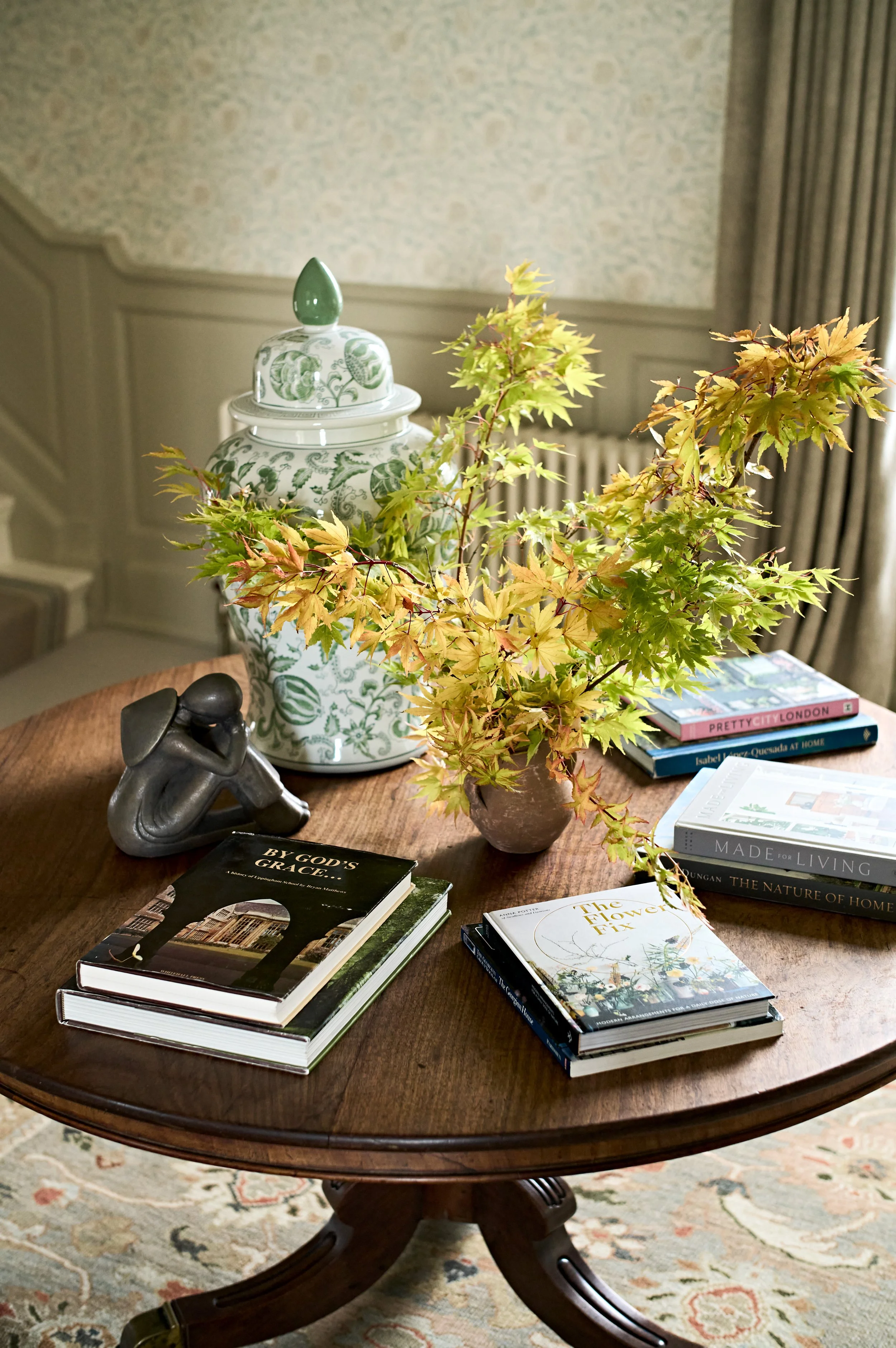 A wooden round table with a decorative ceramic jar, a bronze sculpture of a person, a vase with green and yellow leaves, and several books on interior design, home and garden, and travel. A patterned carpet and curtains are in the background.