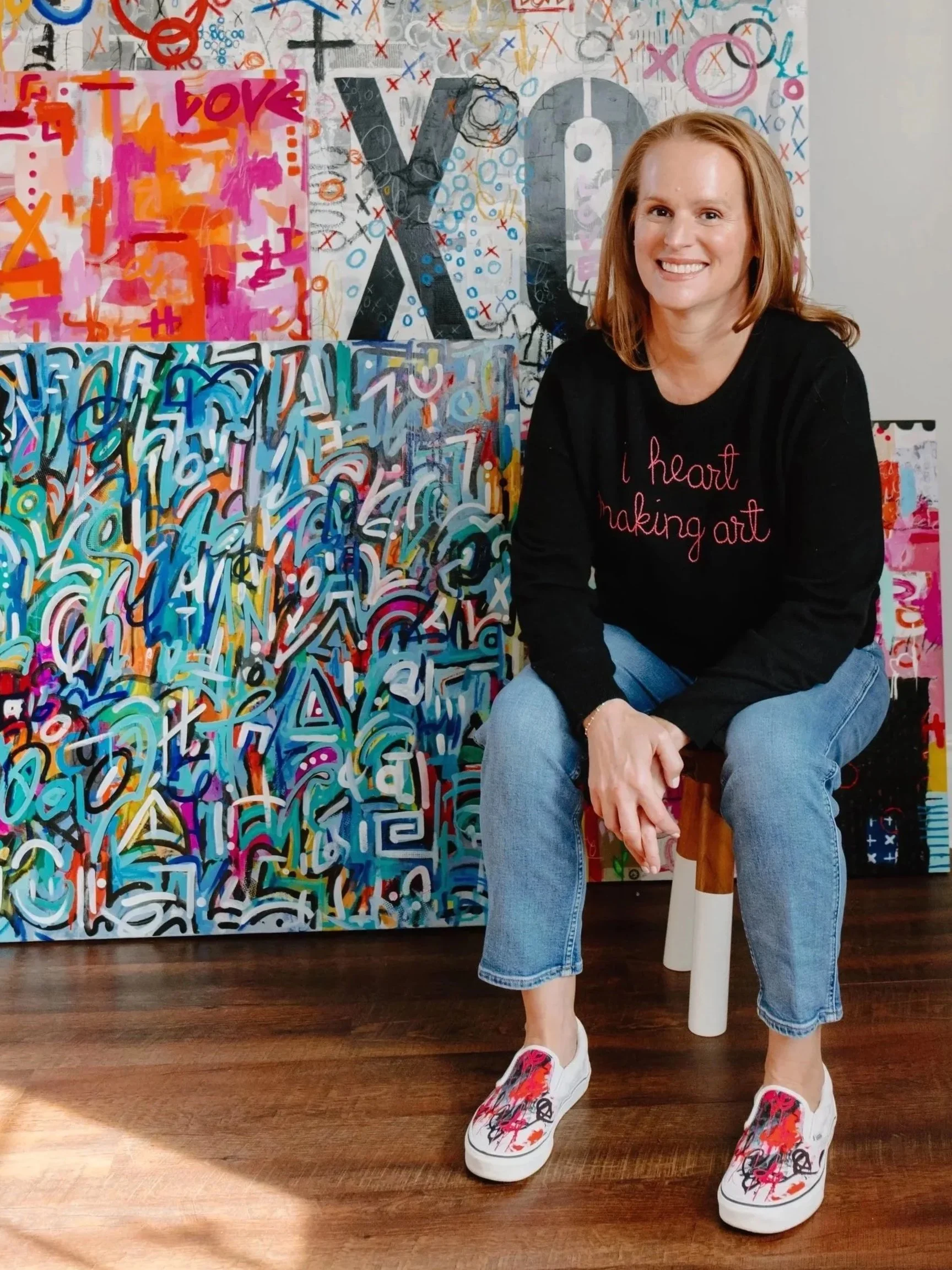 A woman with red hair smiling, sitting on a wooden stool in front of colorful abstract paintings, wearing a black sweatshirt with red lettering, blue jeans, and white sneakers with vibrant paint splatter designs.