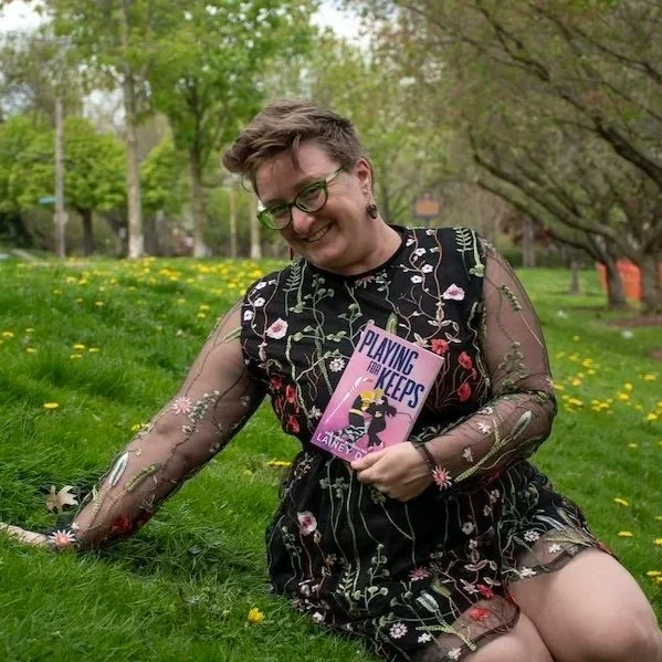 Lainey Davis sits in the grass holding a copy of her book Playing for Keeps