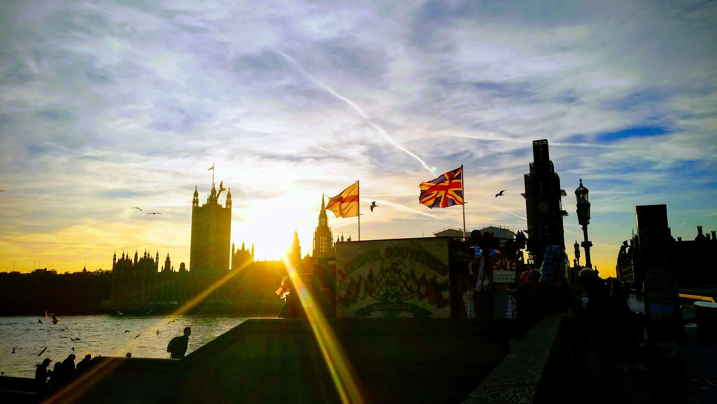 Silhouette de las Casas del Parlamento en Londres con banderas en primer plano durante el atardecer, cielo con nubes y aves volando.