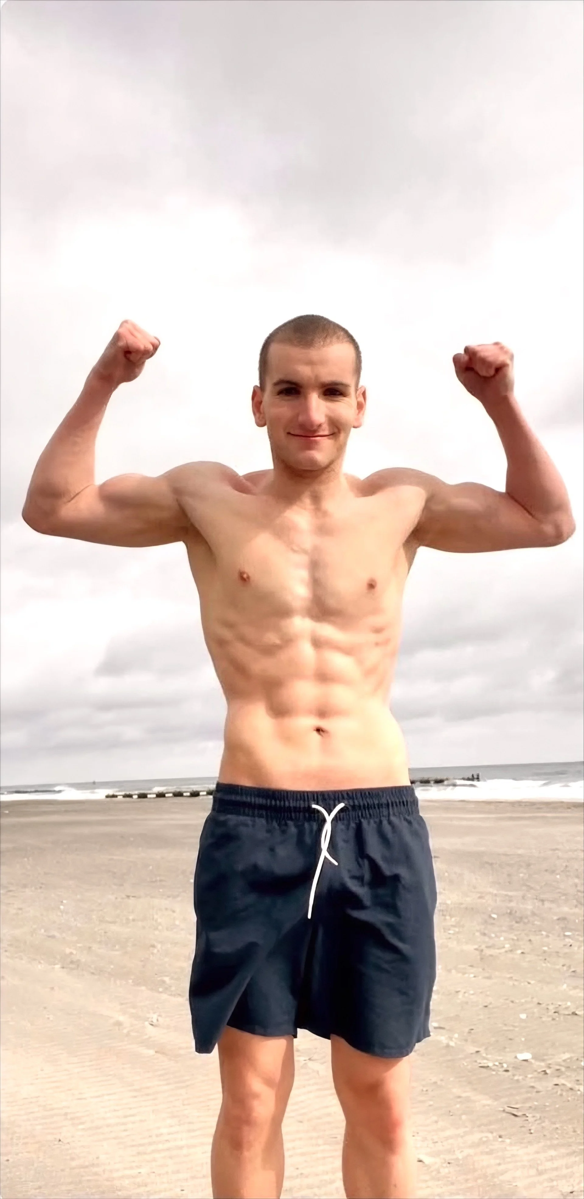 A shirtless young man flexing his biceps and smiling on a beach with an overcast sky in the background.