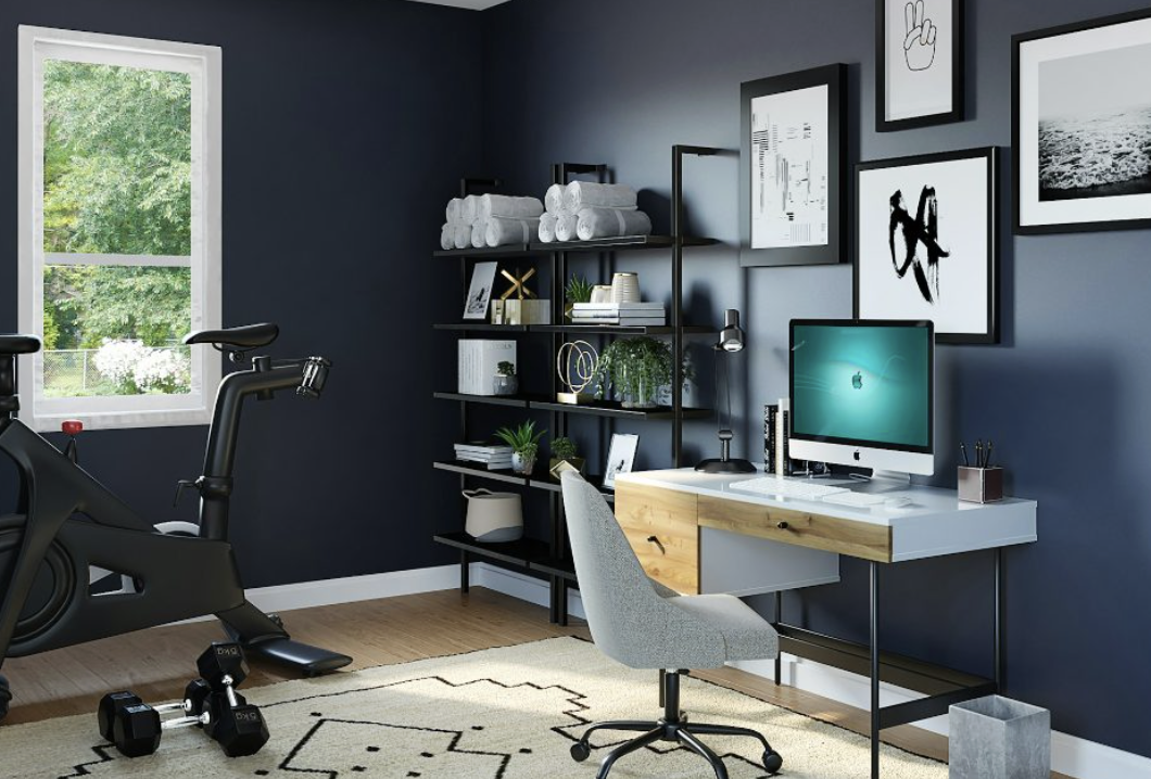 Home office with a black shelving unit, a white desk with a computer, a gray office chair, and a black exercise bike near a window showing greenery outside.
