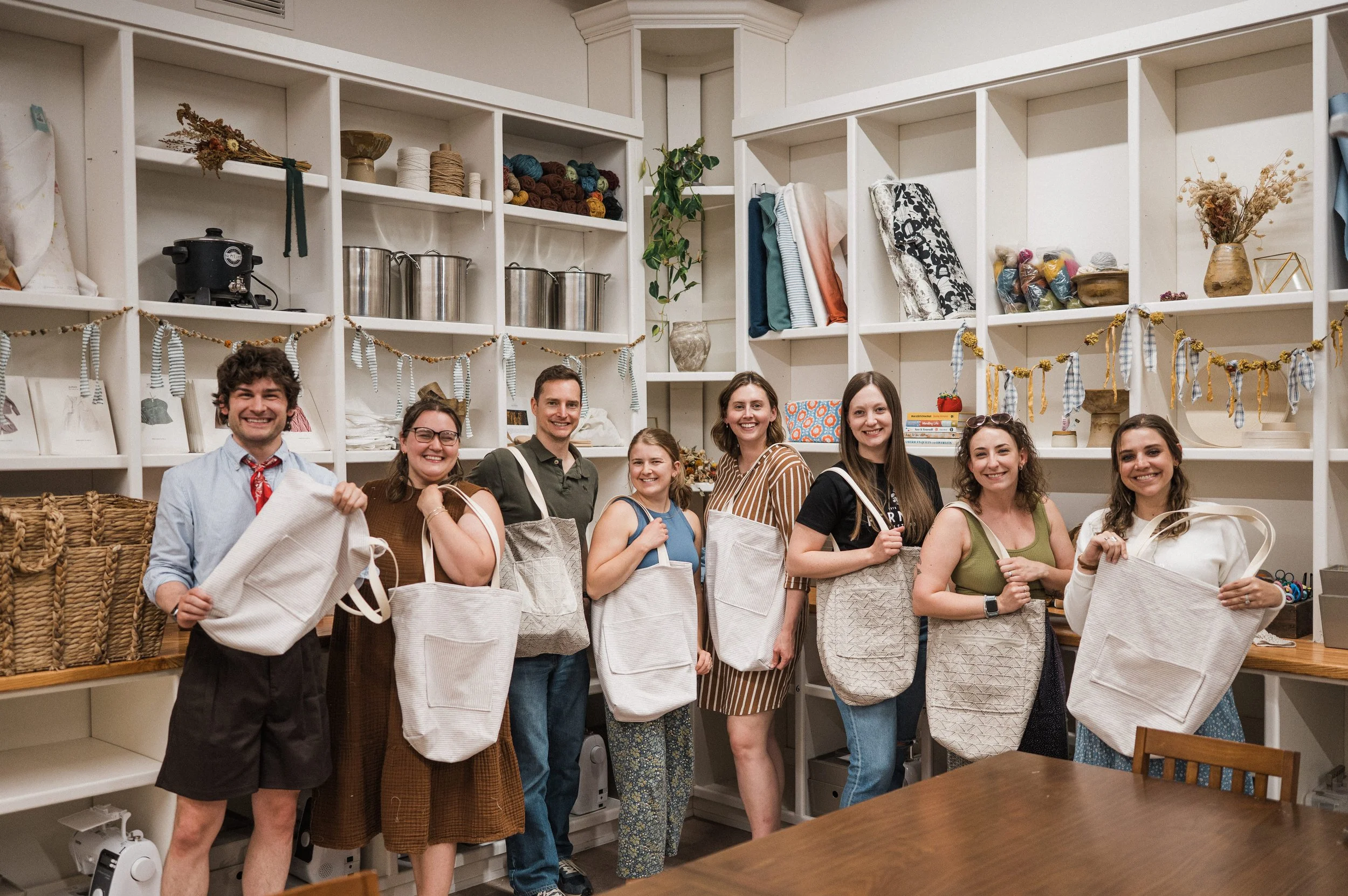 A group of adult men and woman holding tote bags they made in they made in their beginner sewing class
