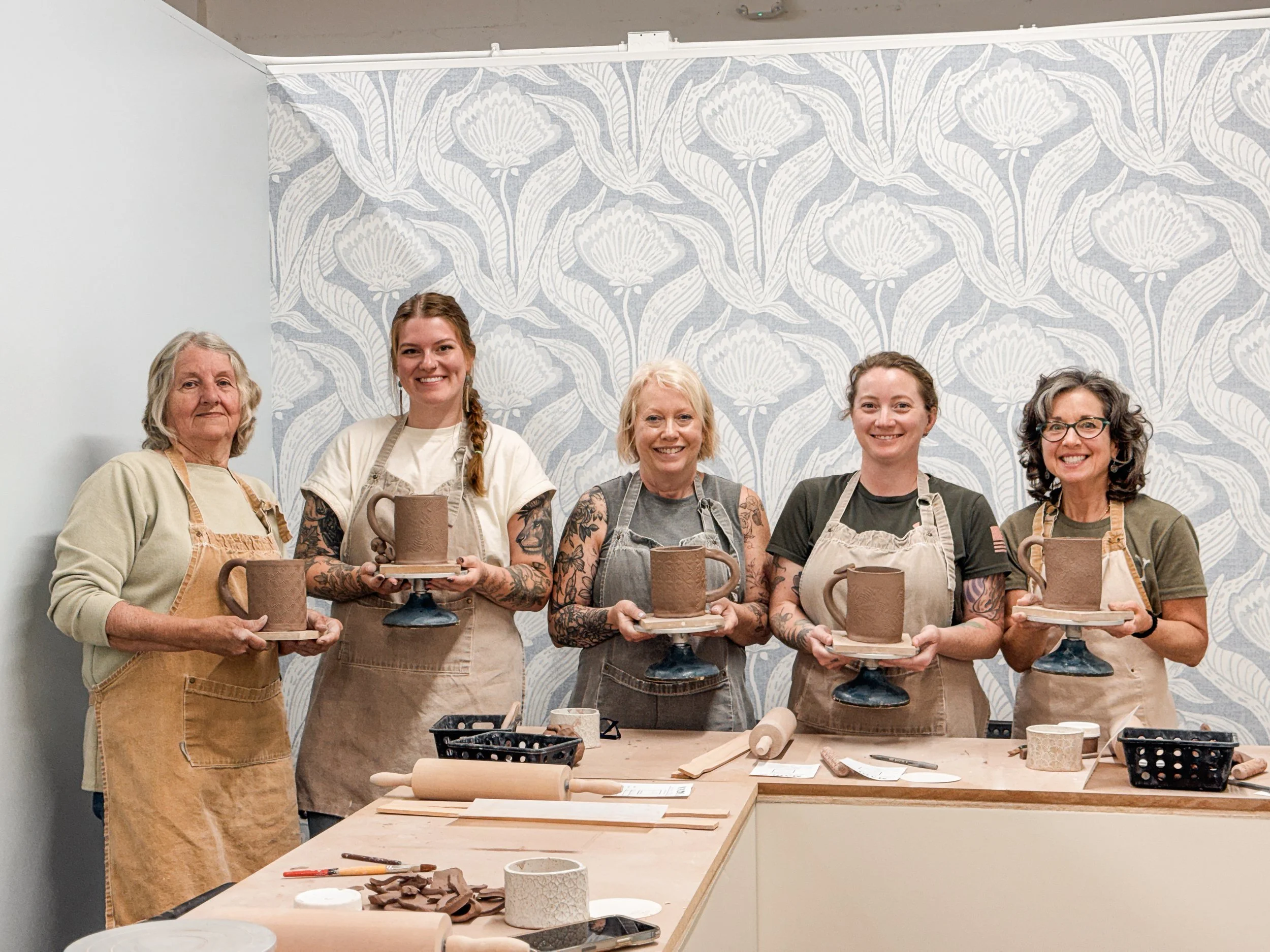 A group of 5 adult woman smiling and holding the mugs they made in a clay workshop experience