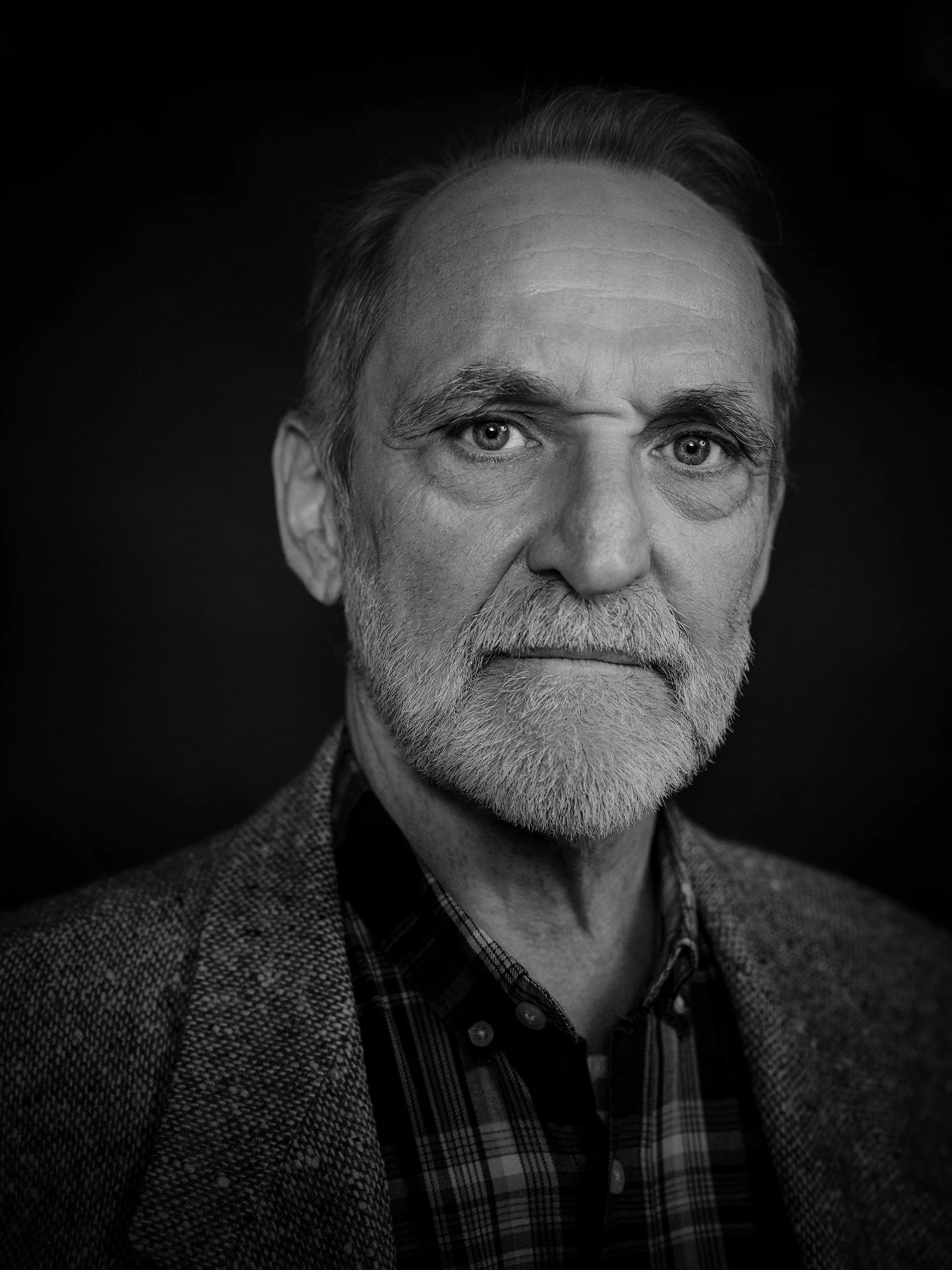 Portrait noir et blanc d’un homme âgé, vu de près, regard direct vers l’objectif. Il porte une barbe grise taillée, une veste en tweed et une chemise à carreaux. La lumière latérale souligne les rides du front et la texture du visage, créant une atmo