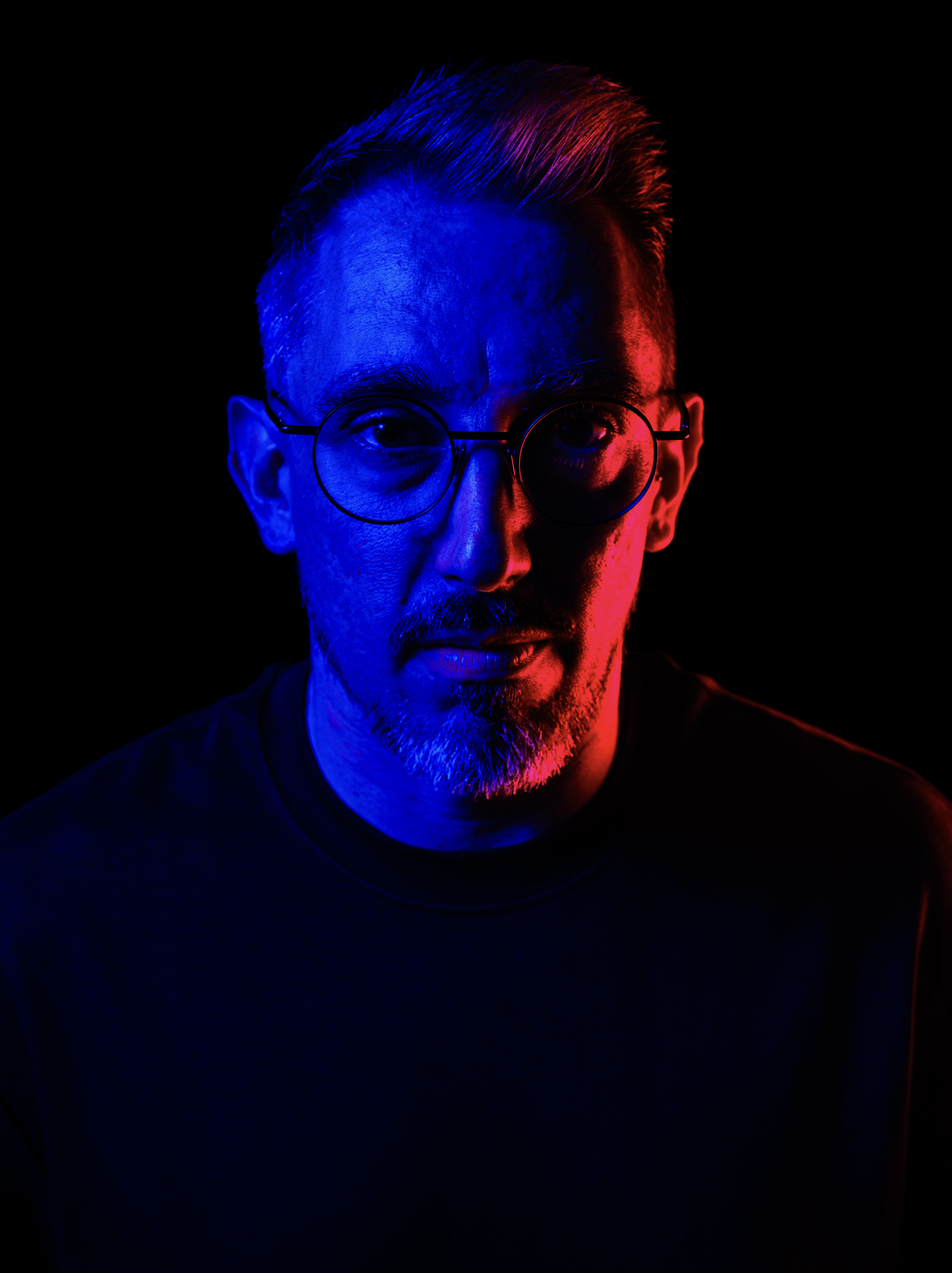 Portrait d'un homme portant des lunettes, illuminé par des lumières rouges et bleues dans un fond noir