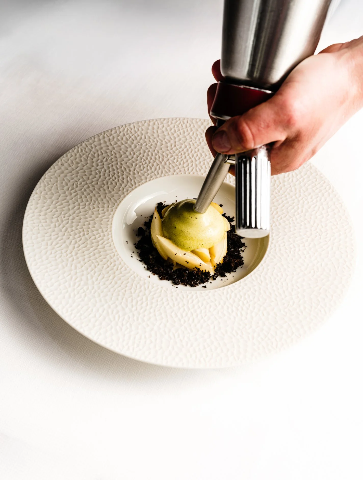 Un dessert élégant avec une boule de glace vert clair sur un lit de biscuits écrasés dans une assiette blanche avec texture. Un instrument sert à ajouter une sauce sur la glace.