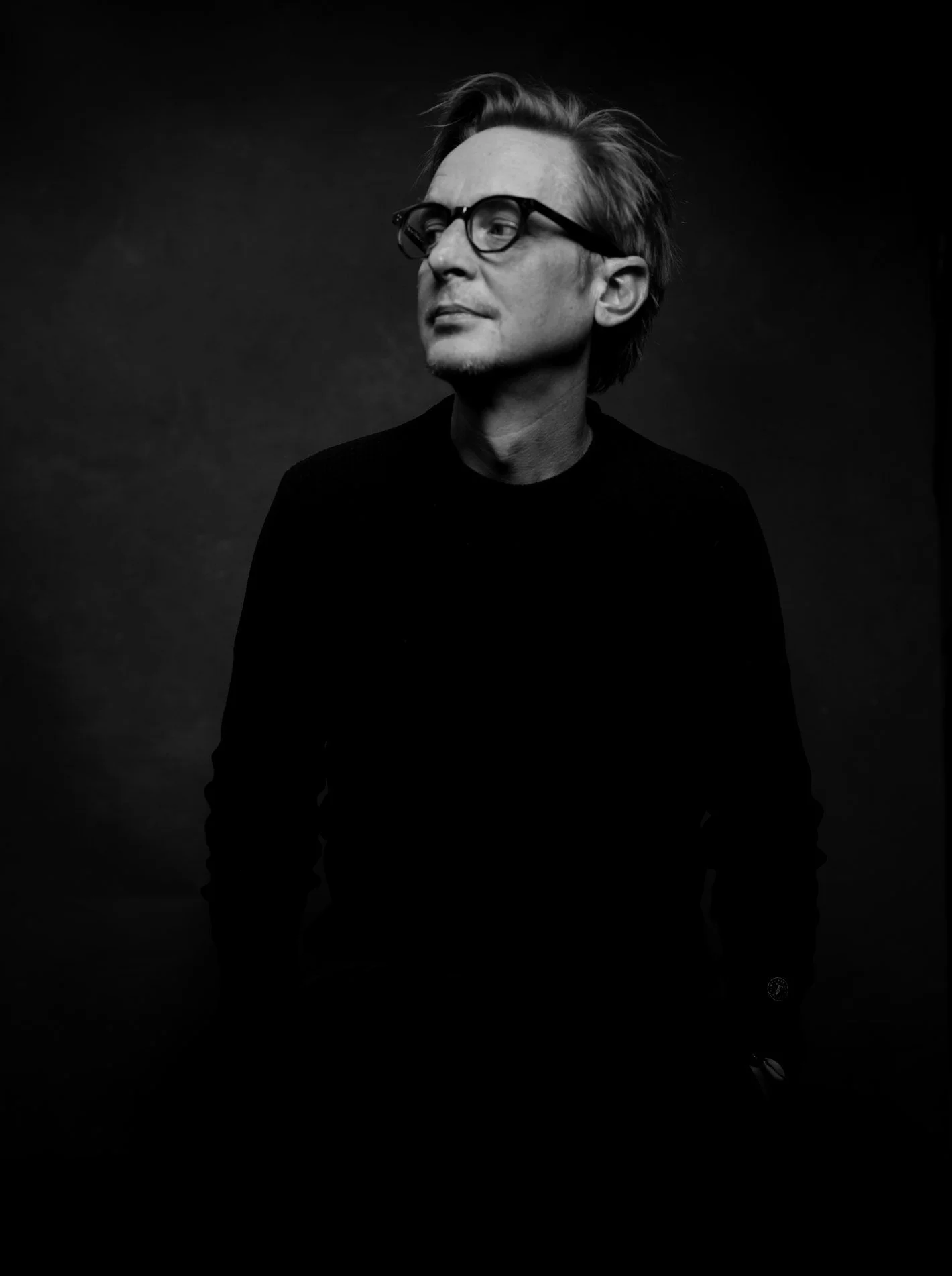 Portrait noir et blanc d'un homme avec des lunettes, cheveux en arrière, regardant vers la gauche, habillé en noir, fond sombre.