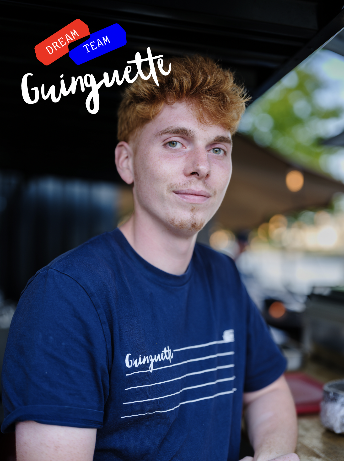 Un jeune homme aux cheveux roux et yeux clairs porte un t-shirt bleu avec l'inscription "Güngutte" en blanc, assis dans un environnement extérieur