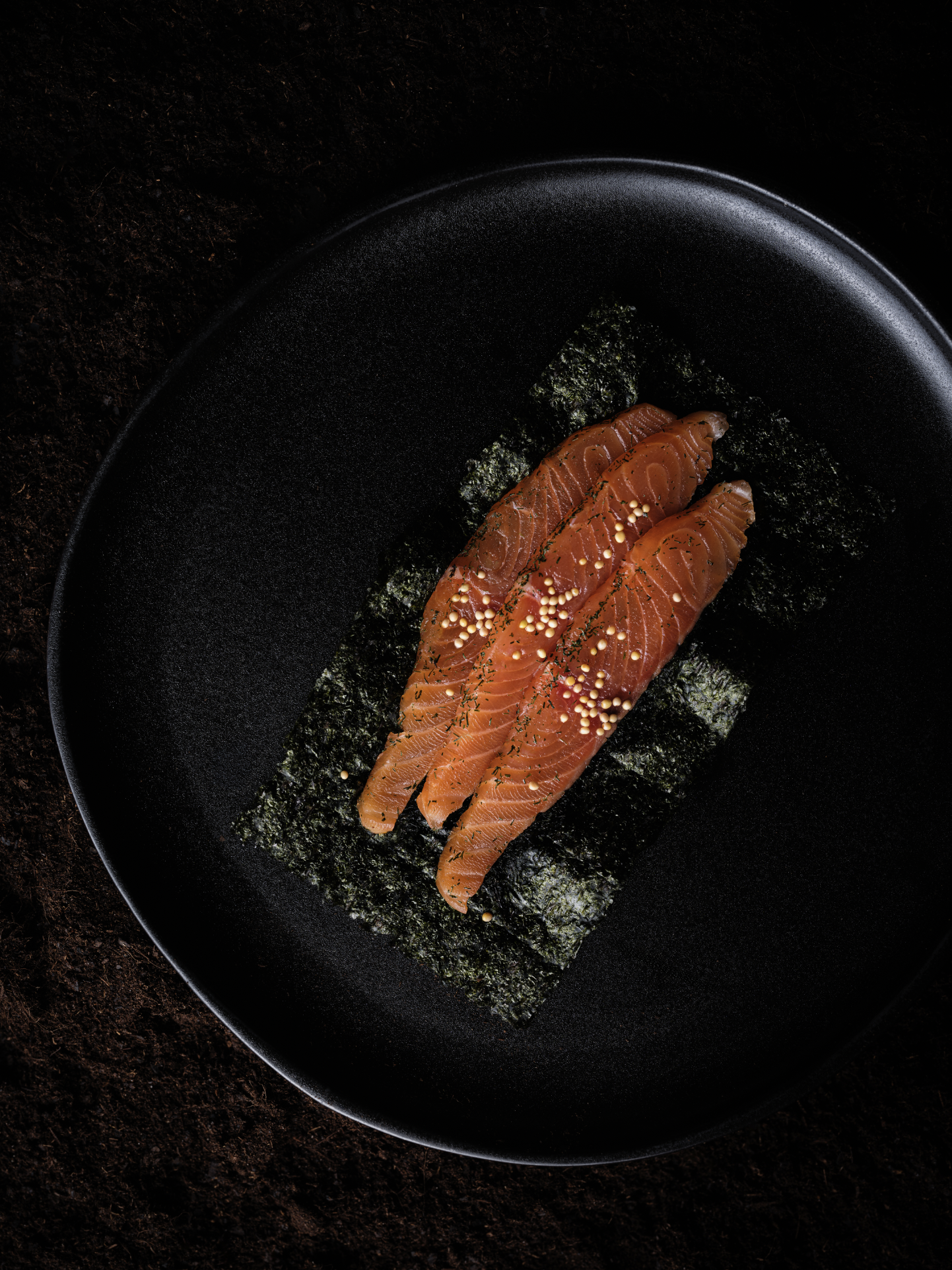Tranches de saumon cru déposées sur une feuille de nori, présentées dans une assiette noire ronde, posée sur un fond de terre sombre. La lumière latérale met en valeur les textures du saumon, du nori et de la surface mate de l’assiette.