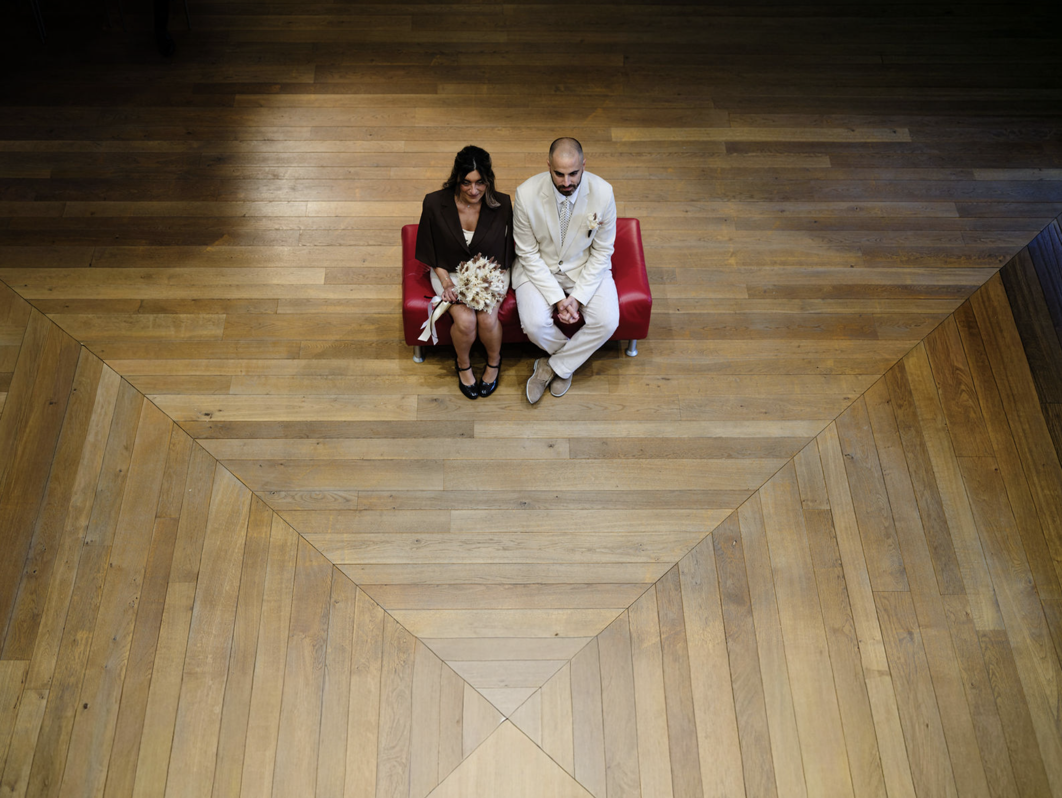 Deux personnes assises sur un banc rouge dans une salle avec un parquet en bois, vues de dessus.