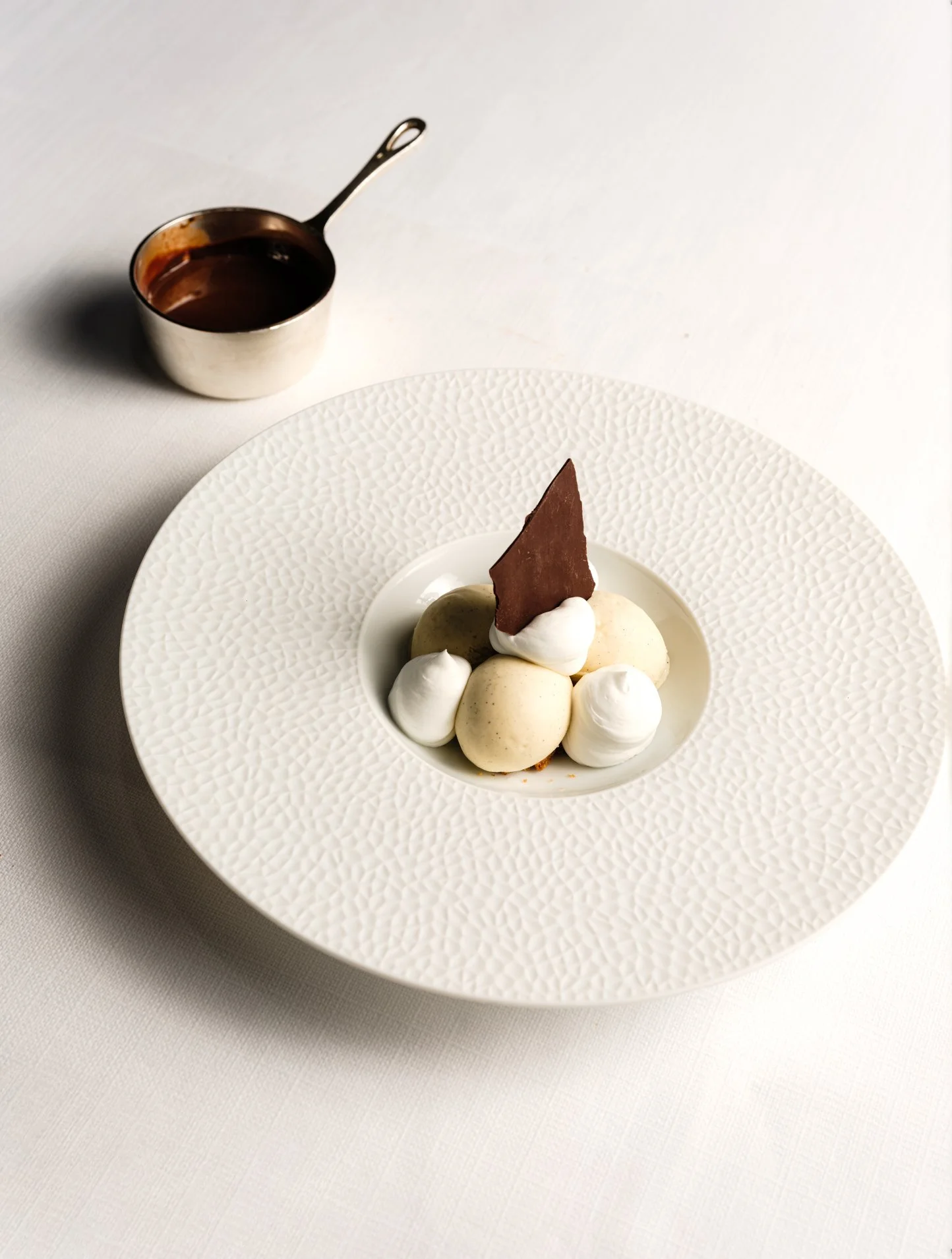 Un dessert avec des boules de glace ou de mousse décorées de crème et d'une fine plaque de chocolat, sur une assiette blanche texturée.