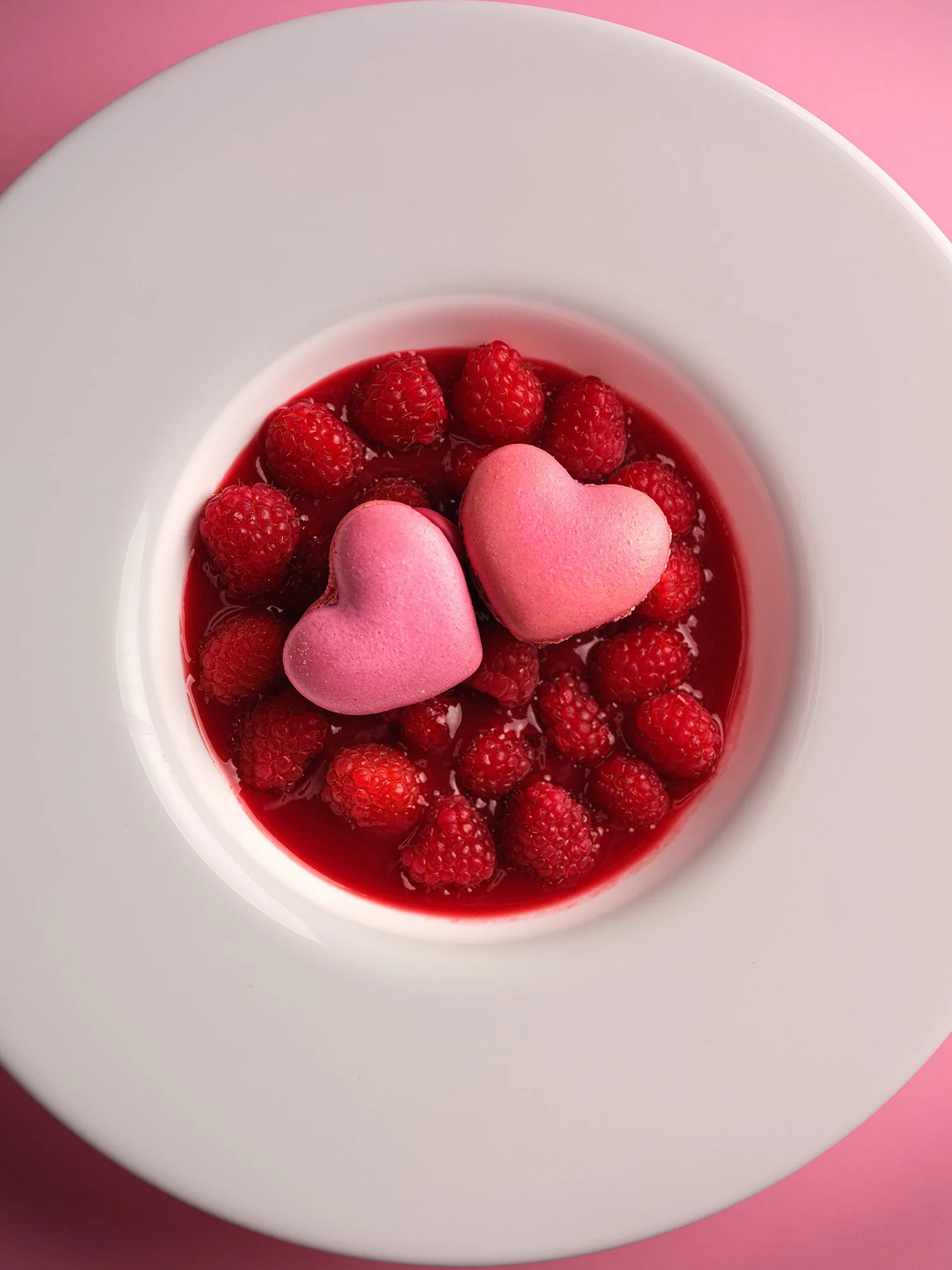 Photographie culinaire minimaliste en vue plongeante : framboises fraîches et deux cœurs rouges disposés dans une assiette blanche sur fond rose, travail de lumière douce et modelé précis.