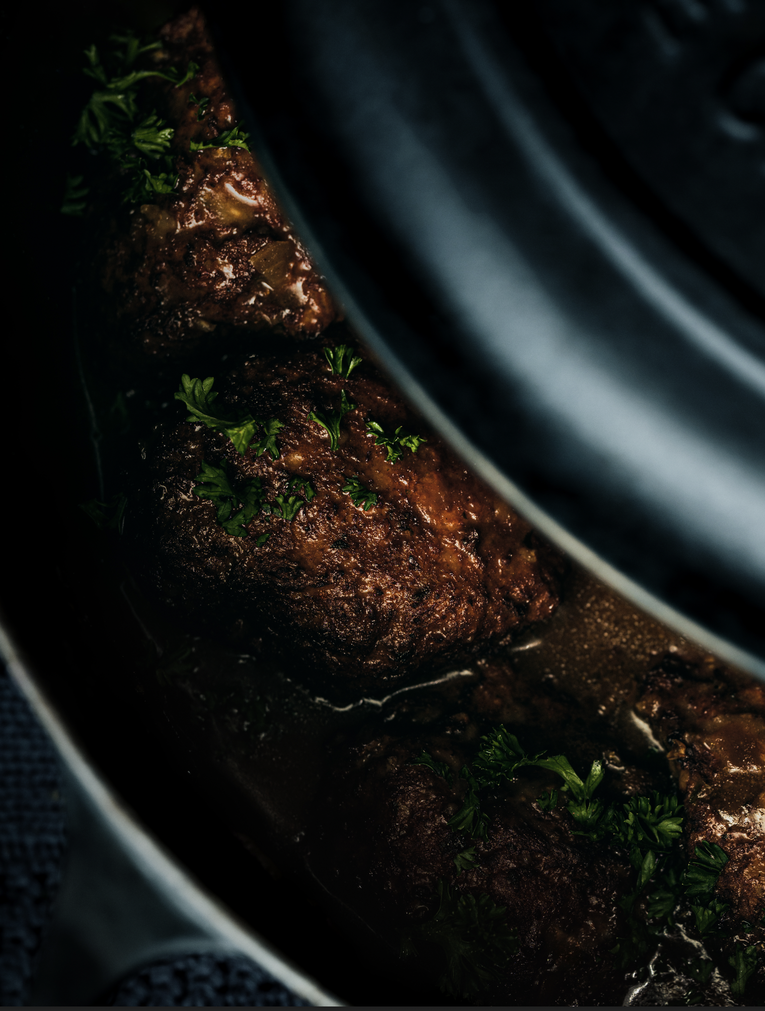 Close-up d’un boulet doré et caramélisé baignant dans une sauce brune, éclairé en clair-obscur, avec quelques feuilles de persil pour la fraîcheur.