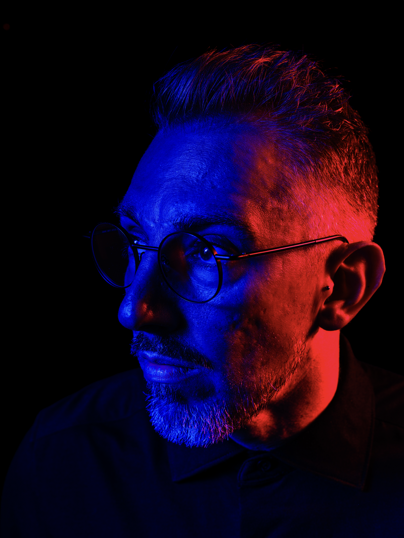 Portrait d'un homme avec des cheveux courts, une barbe et des lunettes rondes, éclairé par des lumières rouge et bleue sur un fond noir.