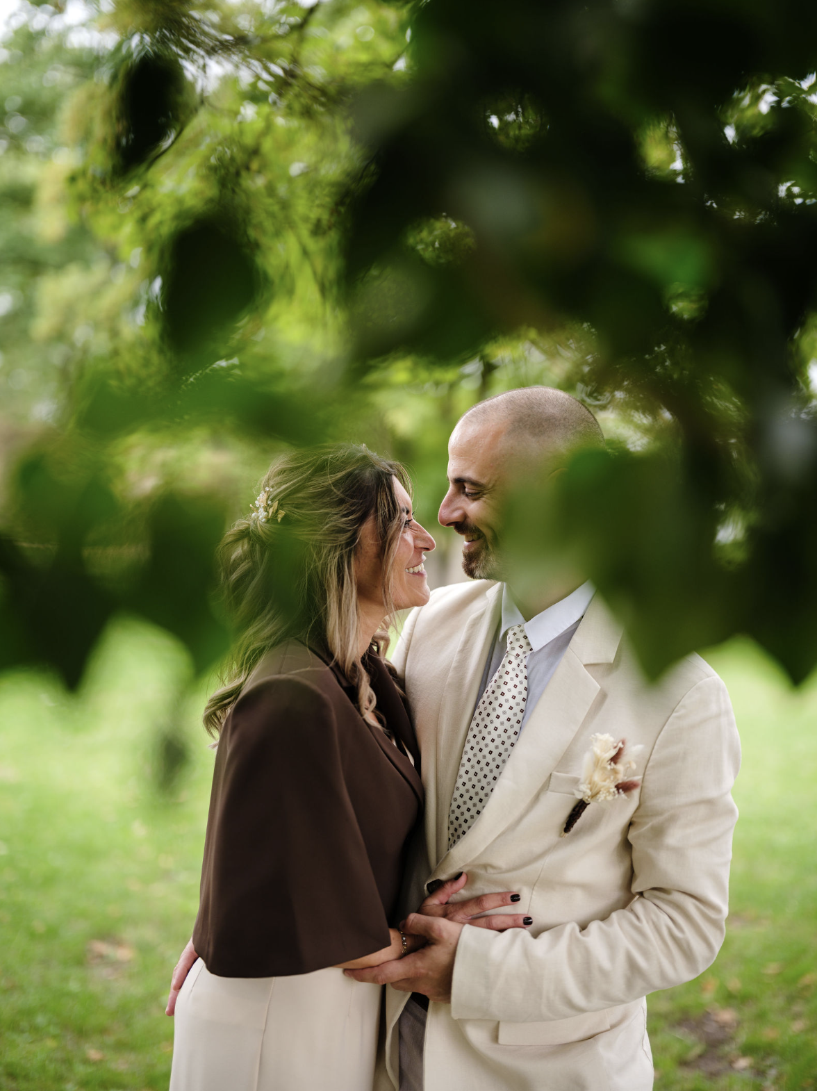 Un couple souriant, habillé formellement, s'enlace dans un parc verdoyant, encadré par des feuilles d'arbres.