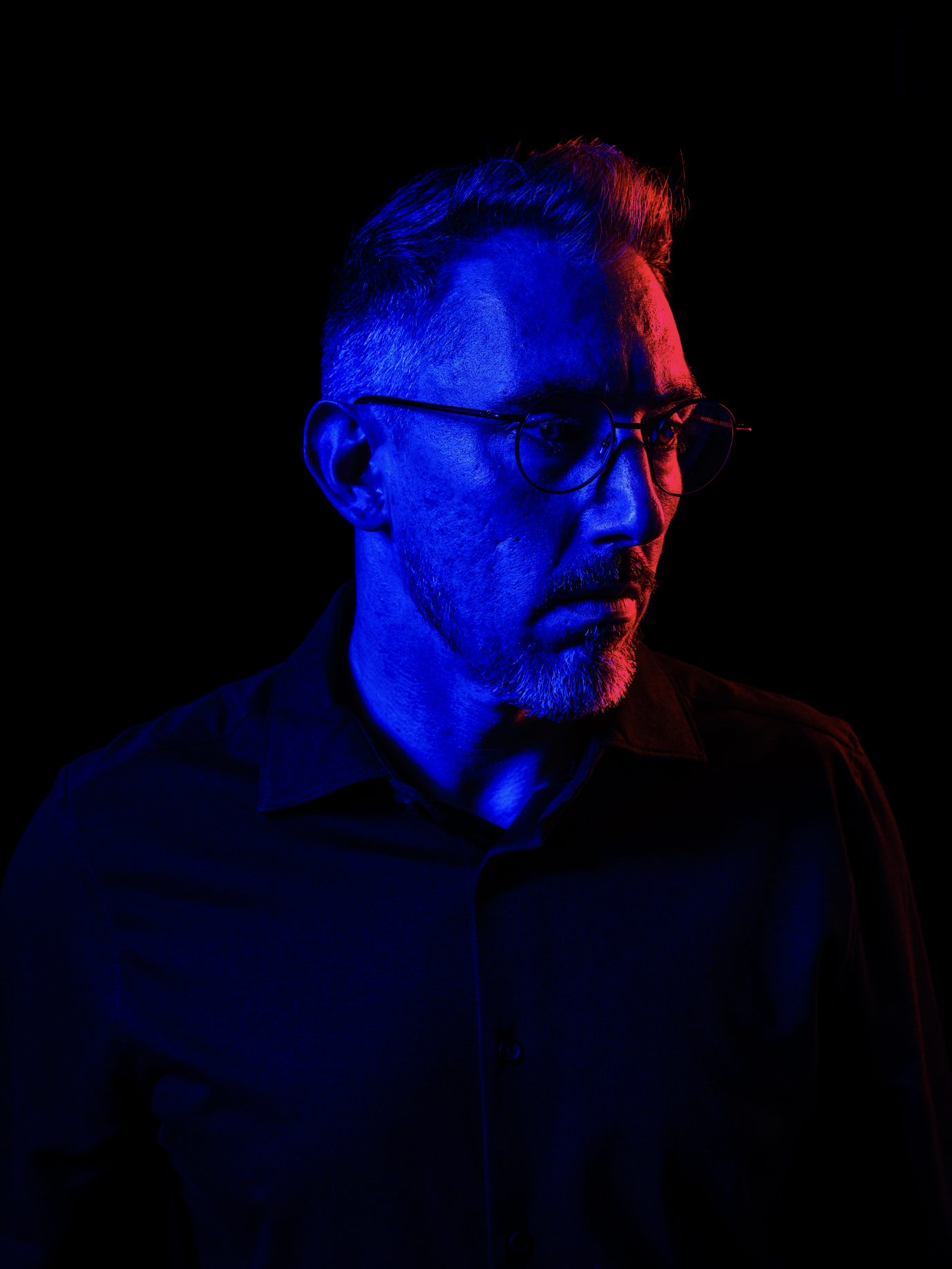 Portrait d'un homme avec des cheveux gris, portant des lunettes rondes, éclairé par une lumière bleue et rouge sur un fond noir.
