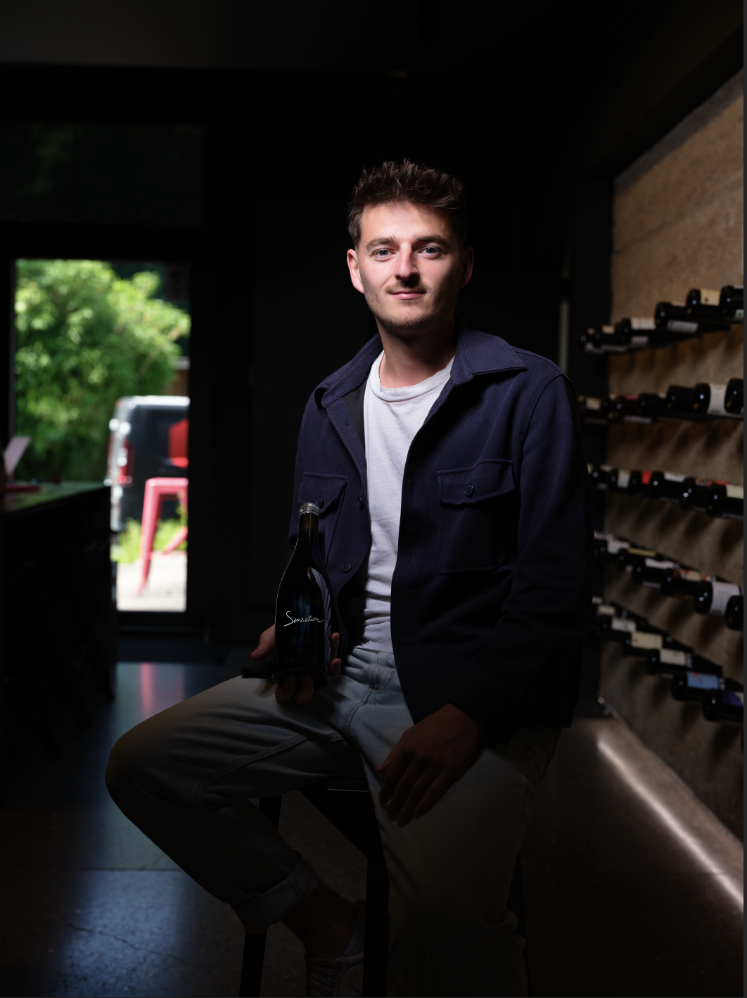 Un jeune homme assis sur un tabouret dans une cave à vin moderne, éclairé par une lumière naturelle provenant d'une porte ouverte à l'arrière. Il tient une bouteille de vin sombre étiquetée "Sensation" et sourit légèrement face à l'objectif. Des bout
