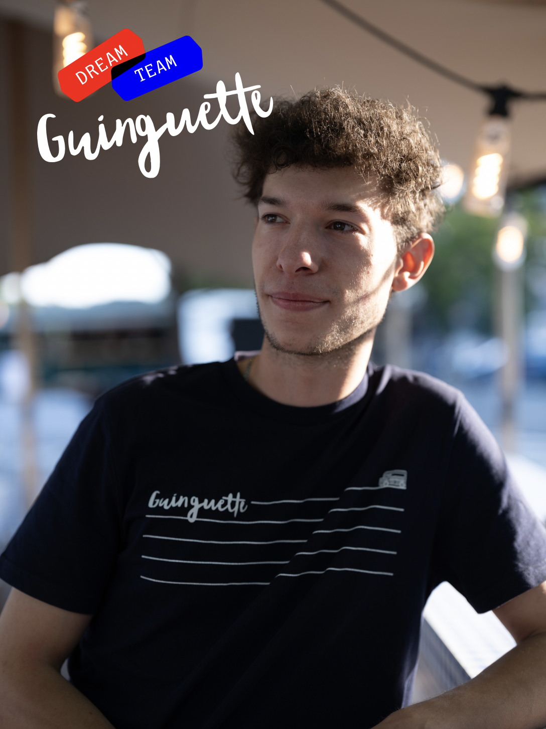 Jeune homme avec cheveux bouclés portant un t-shirt noir avec l'inscription "Guinguette" sur fond flou de lumières et de décor extérieur.